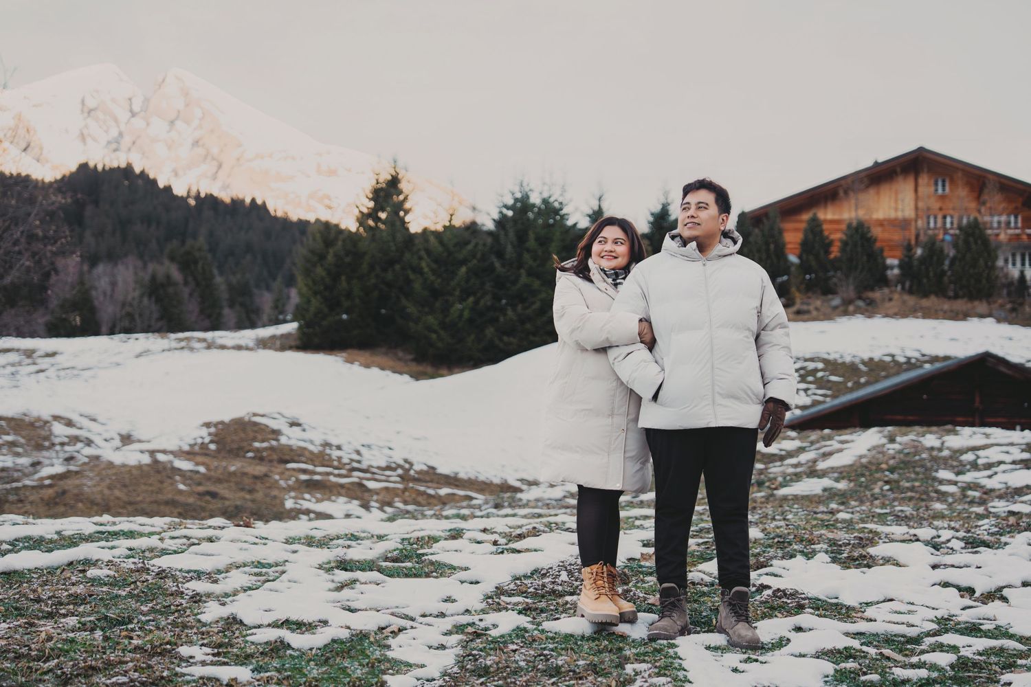 Couple in white winter jackets standing together on snowy alpine meadow at dusk.