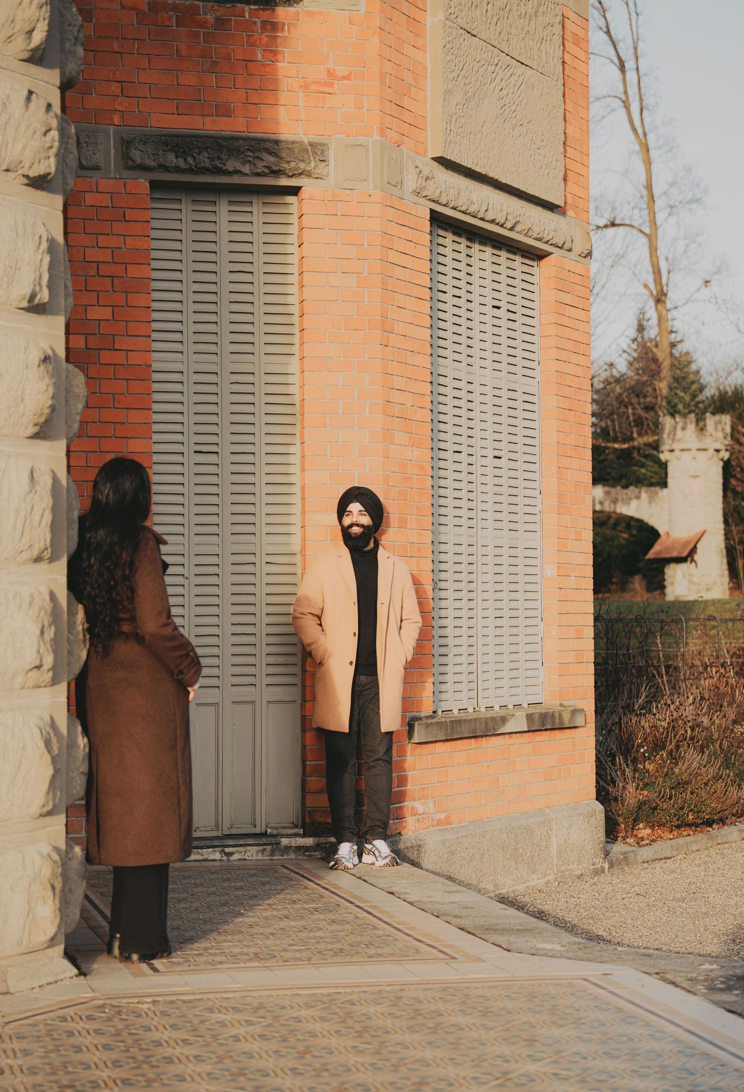 Man in turban and beige coat standing by a brick building with shuttered windows outdoors.