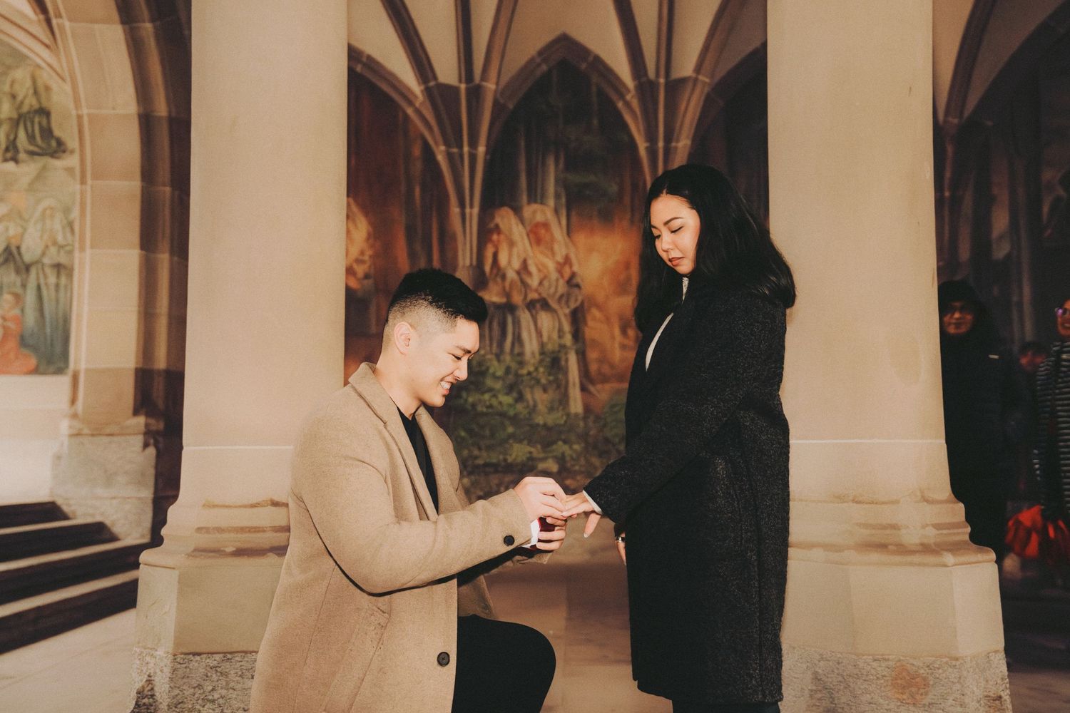 Man presenting engagement ring while kneeling before woman in gothic arcade with fresco murals.
