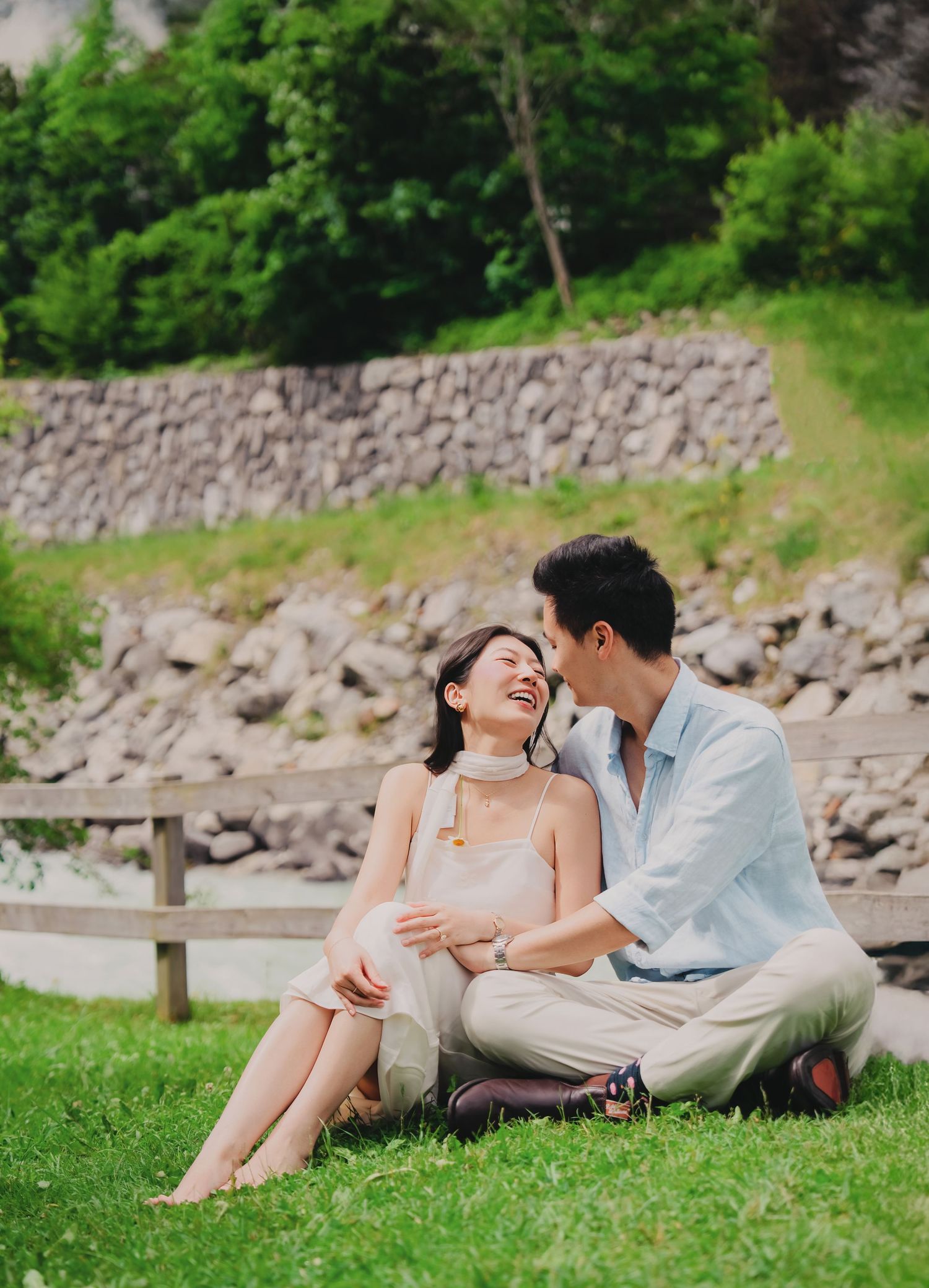 Happy couple sitting on grass laughing together near a stone wall in a scenic green setting.