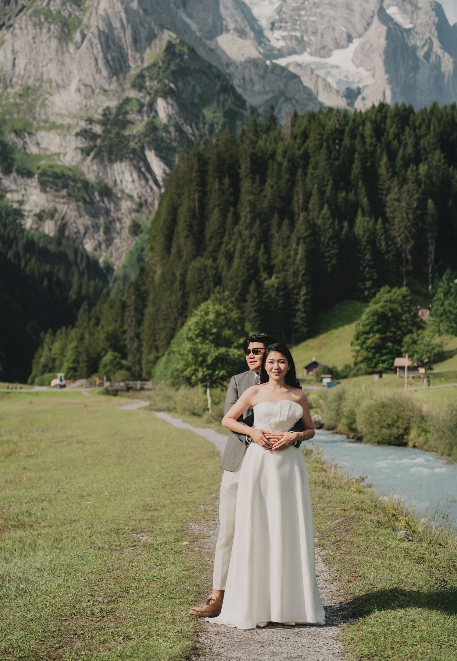 Groom embracing bride from behind beside a turquoise alpine stream with forested mountains in the background.
