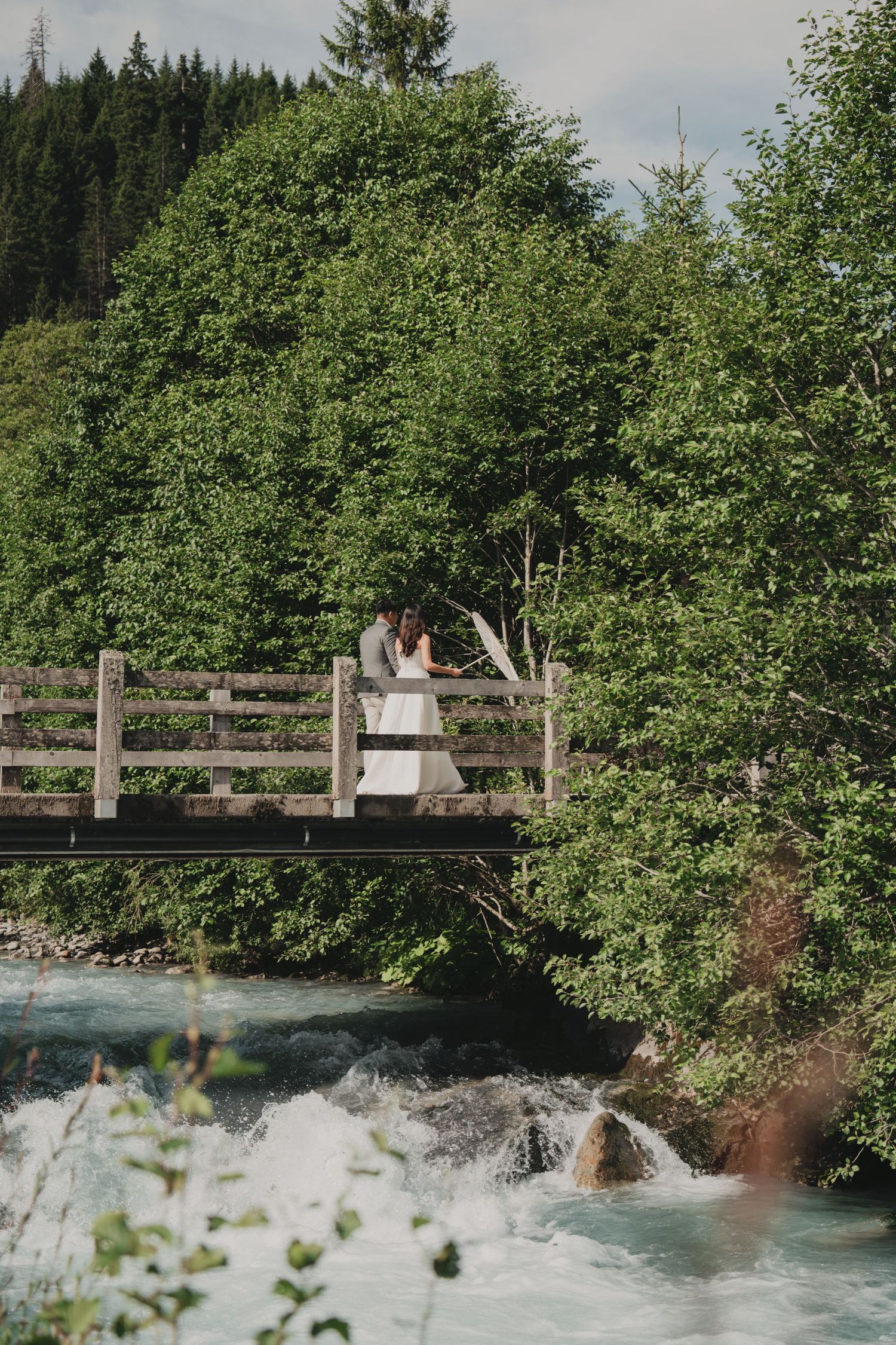 Bride and groom share intimate moment on wooden bridge over rushing alpine river surrounded by lush trees.
