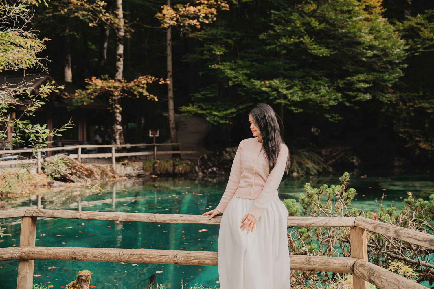 Elegant woman in white skirt and lace top gazing down by a wooden fence near a turquoise lake.