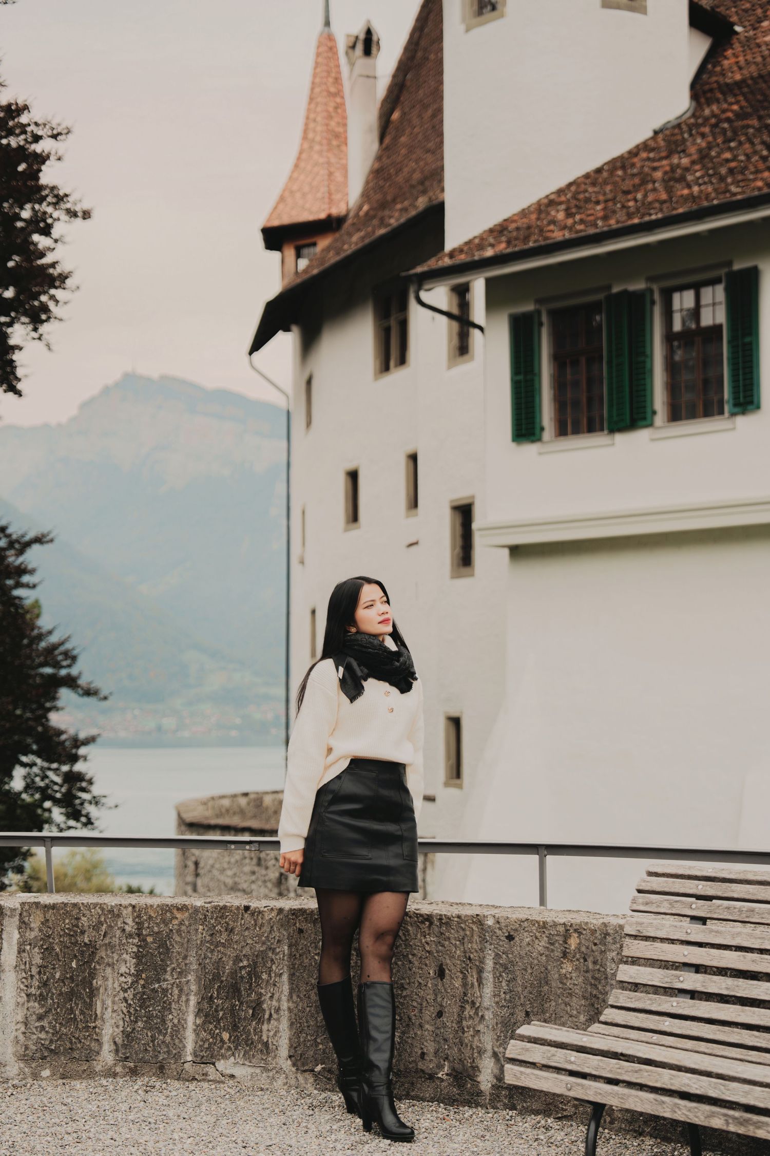 Woman in cream sweater and black leather skirt poses near white Swiss castle with lake backdrop.