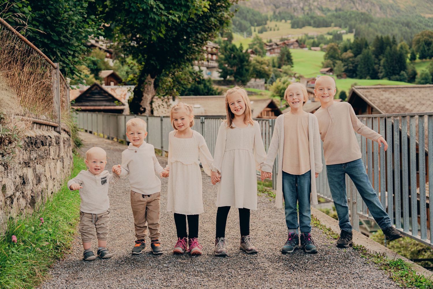 Six young blonde children holding hands in a line on a gravel path with Alpine village scenery behind them.