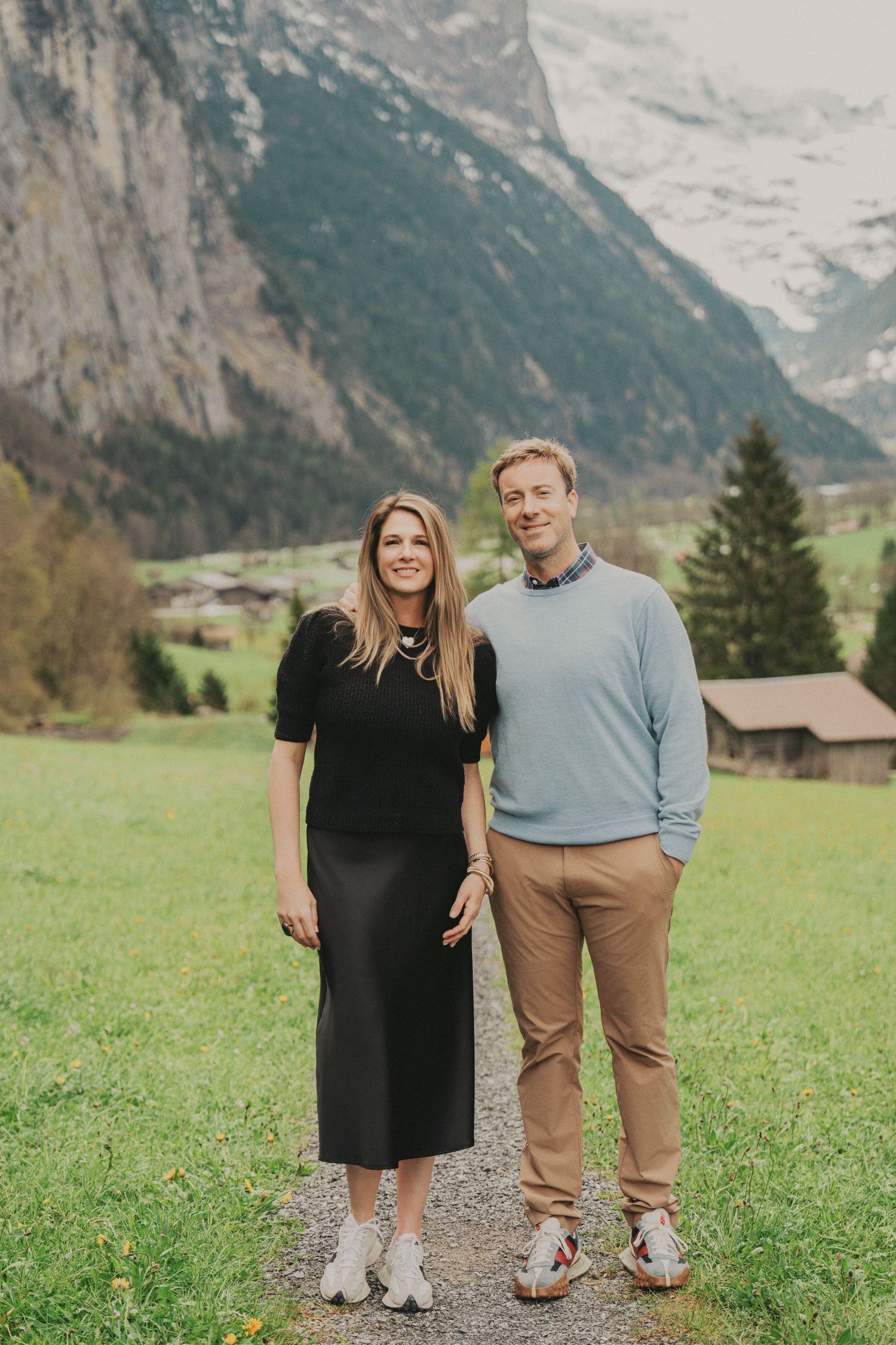 Stylish couple posing on path in Swiss alpine valley with dramatic mountain cliffs in background.