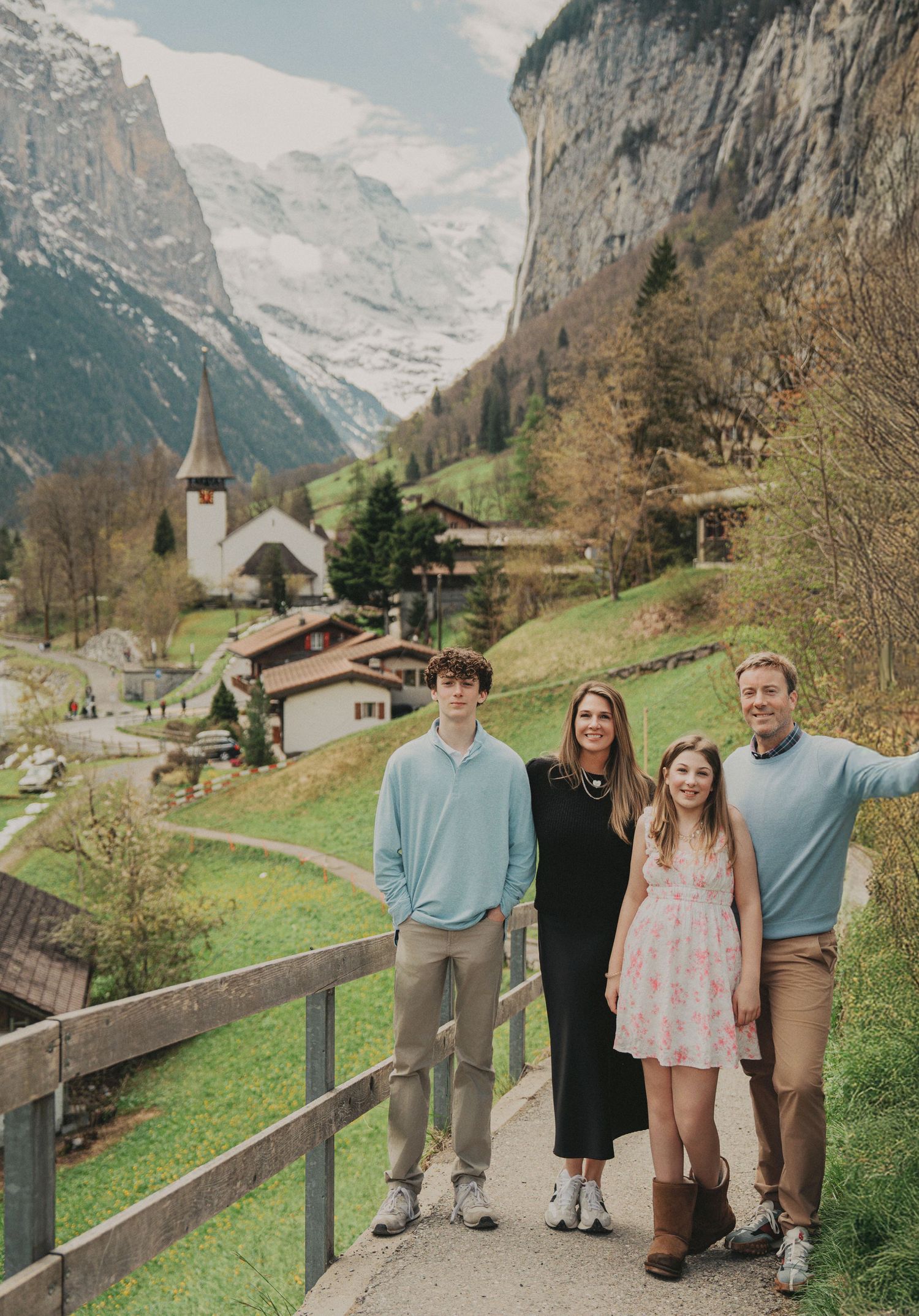Family of four smiling together on a scenic mountain path in a picturesque Swiss village with a church and waterfall.