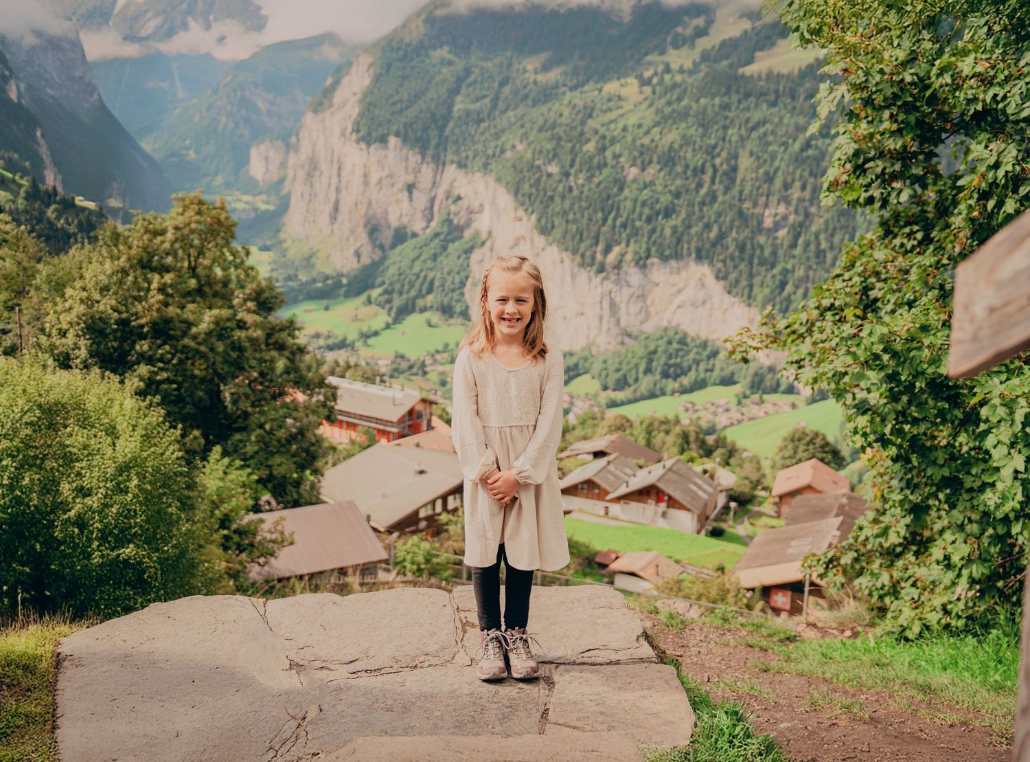 Smiling blonde girl in cream dress standing on stone platform with picturesque Swiss mountain village backdrop.