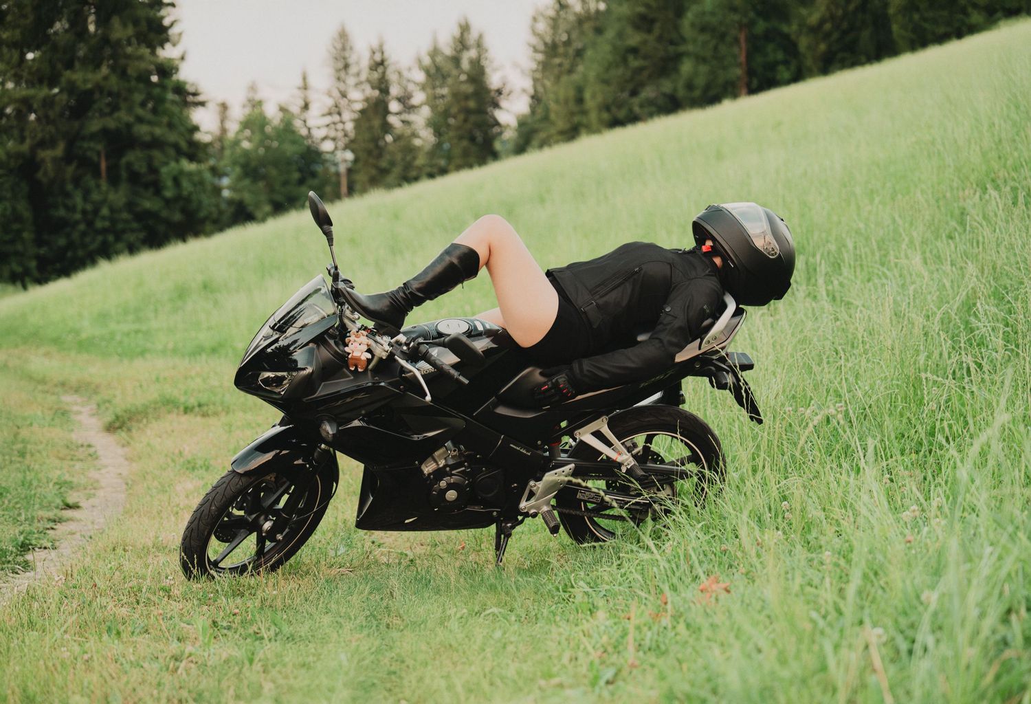 Person lying across a black sport motorcycle on a grassy hillside trail with forest in background.