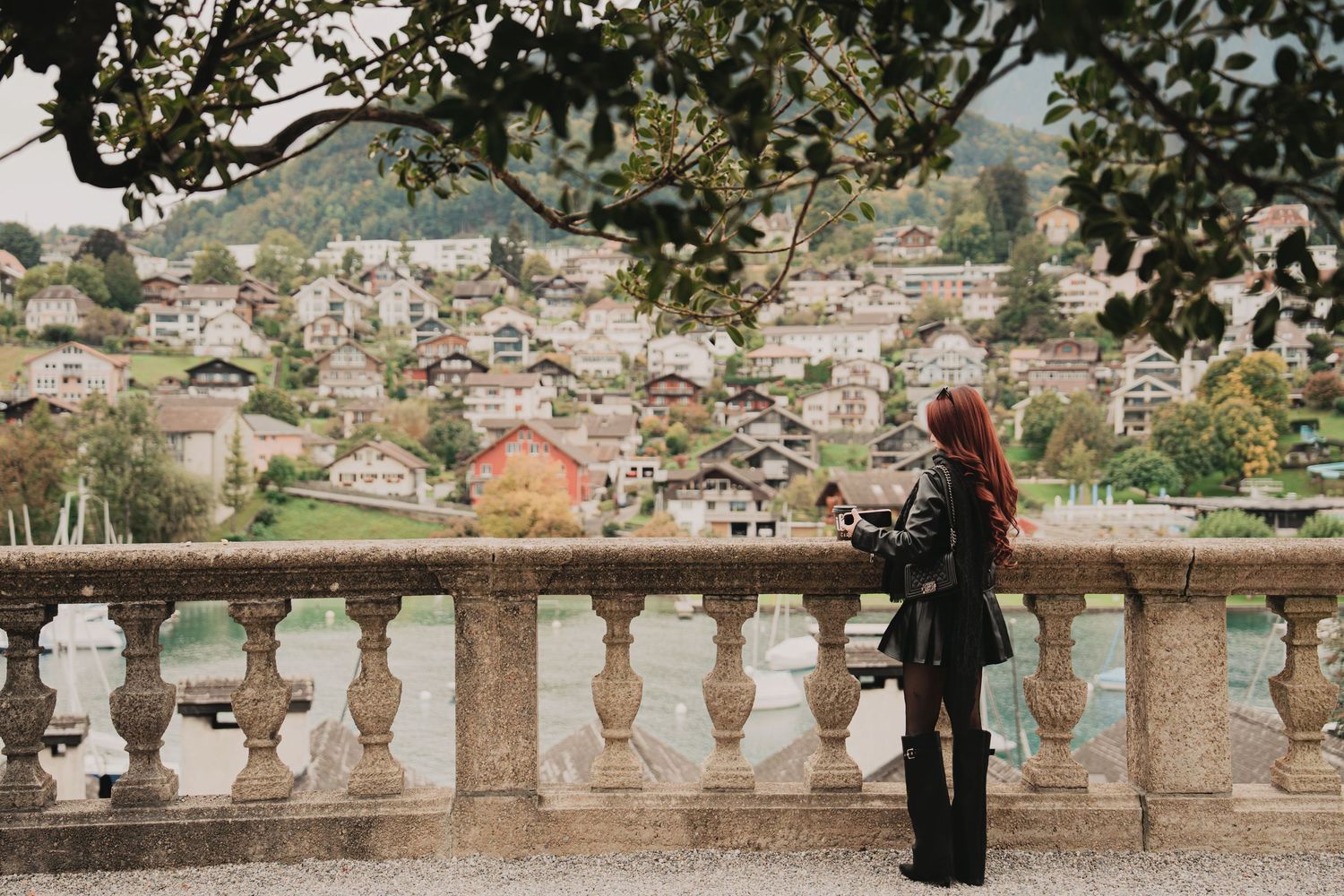 Woman with vibrant red hair stands at ornate stone railing photographing scenic Swiss alpine village.