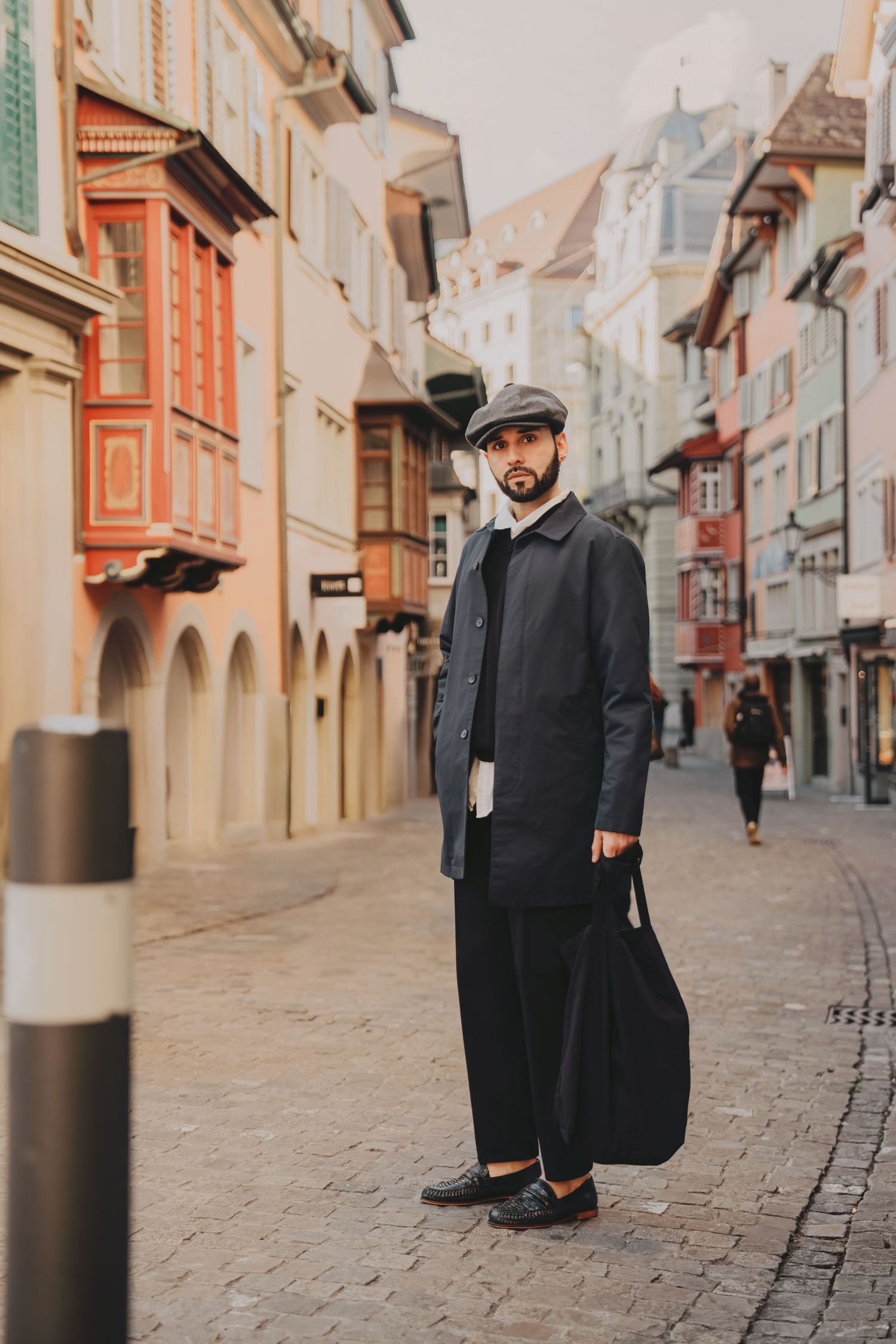 Bearded man in dark coat and newsboy cap holding bag while standing on picturesque European cobblestone street.
