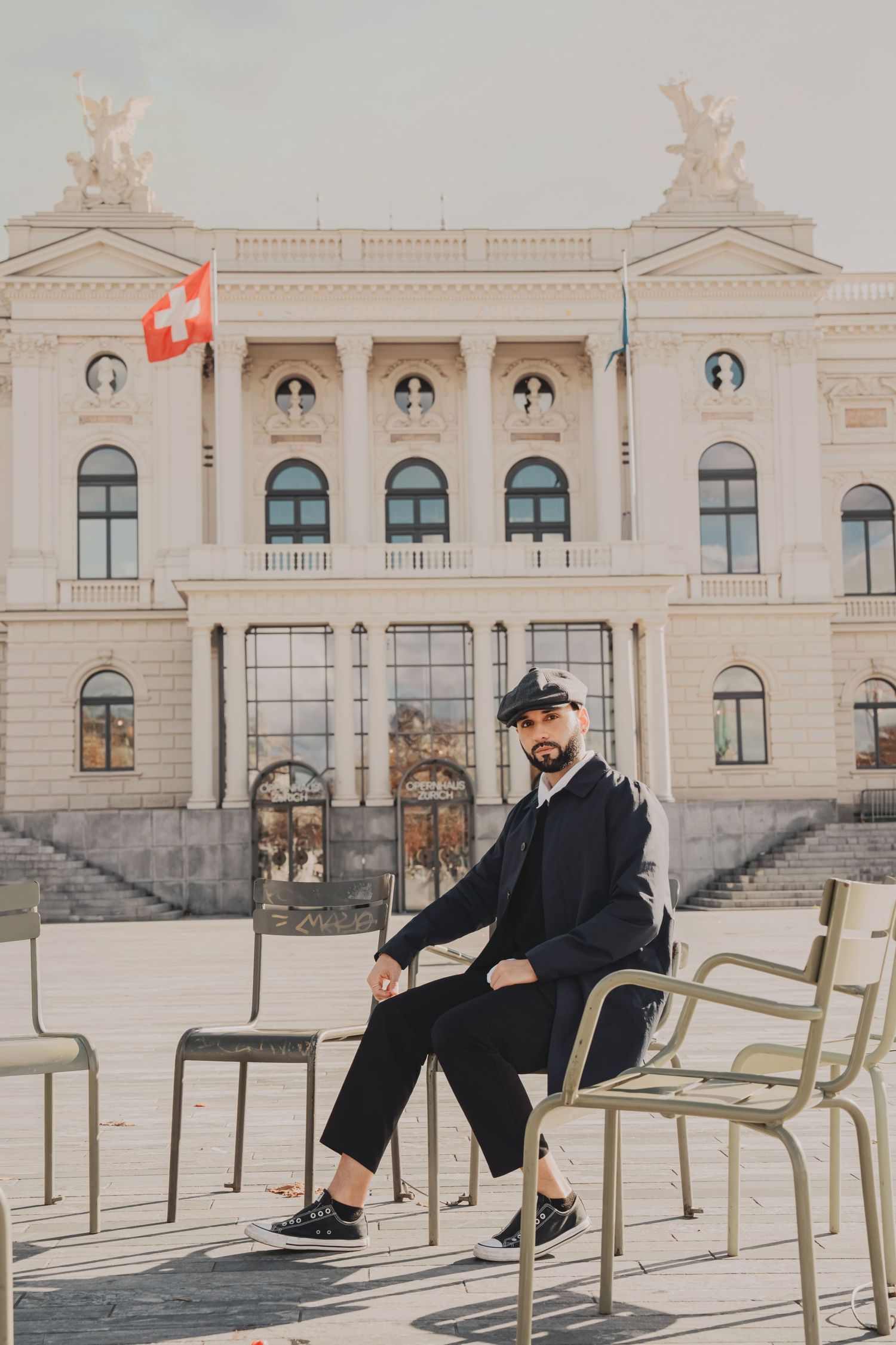 Stylish man in flat cap sitting before grand neoclassical building with Swiss flag in background.