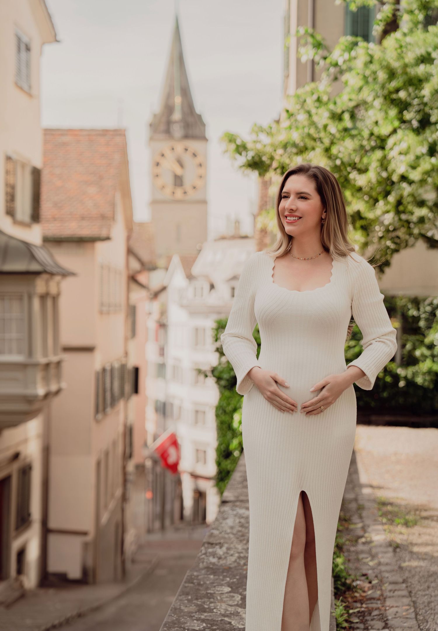 Smiling pregnant woman in fitted white dress cradles baby bump on cobblestone street in Zurich old town.