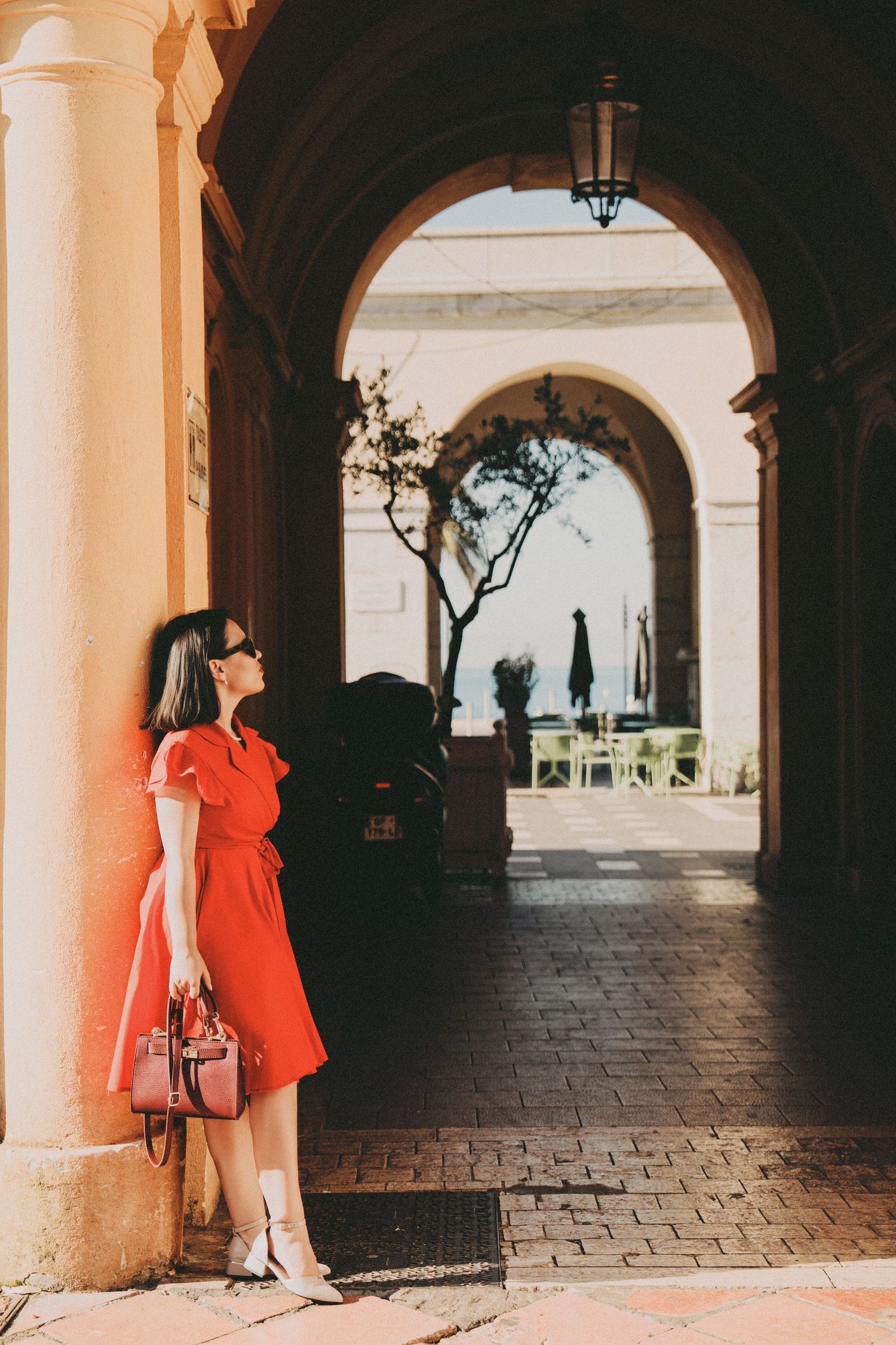 Woman in red wrap dress leaning against column in elegant arched arcade in Monaco.