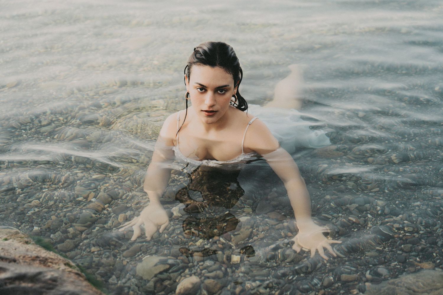 Woman in wet white bikini top emerging from shallow rocky water, looking directly at camera.