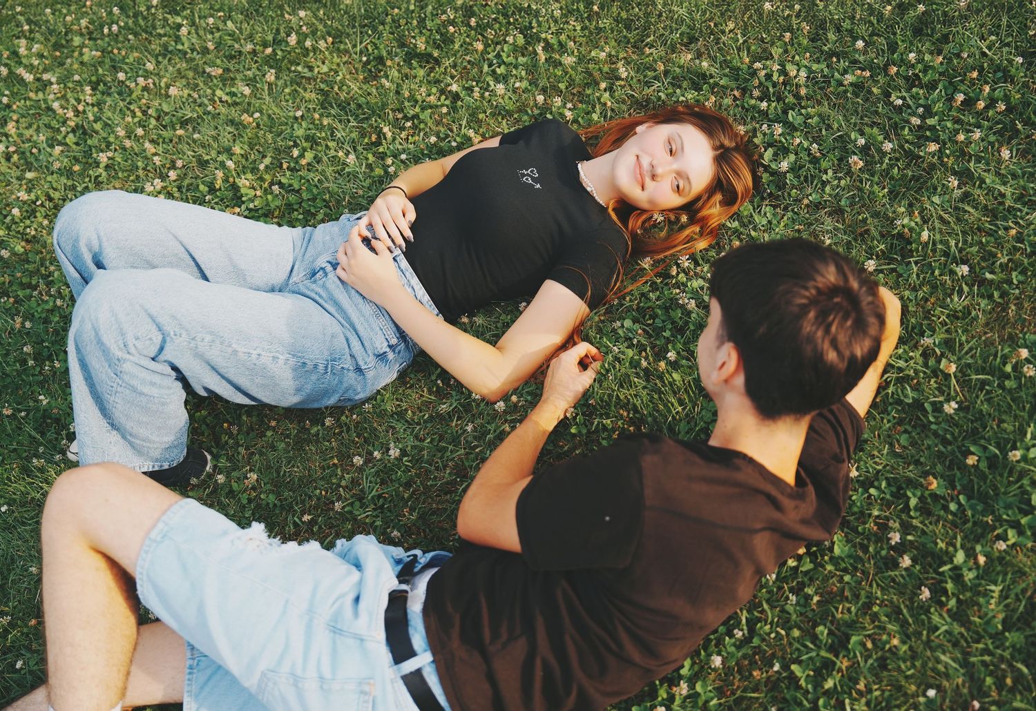 Young couple relaxing on green grass, woman with red hair lying down smiling at partner.