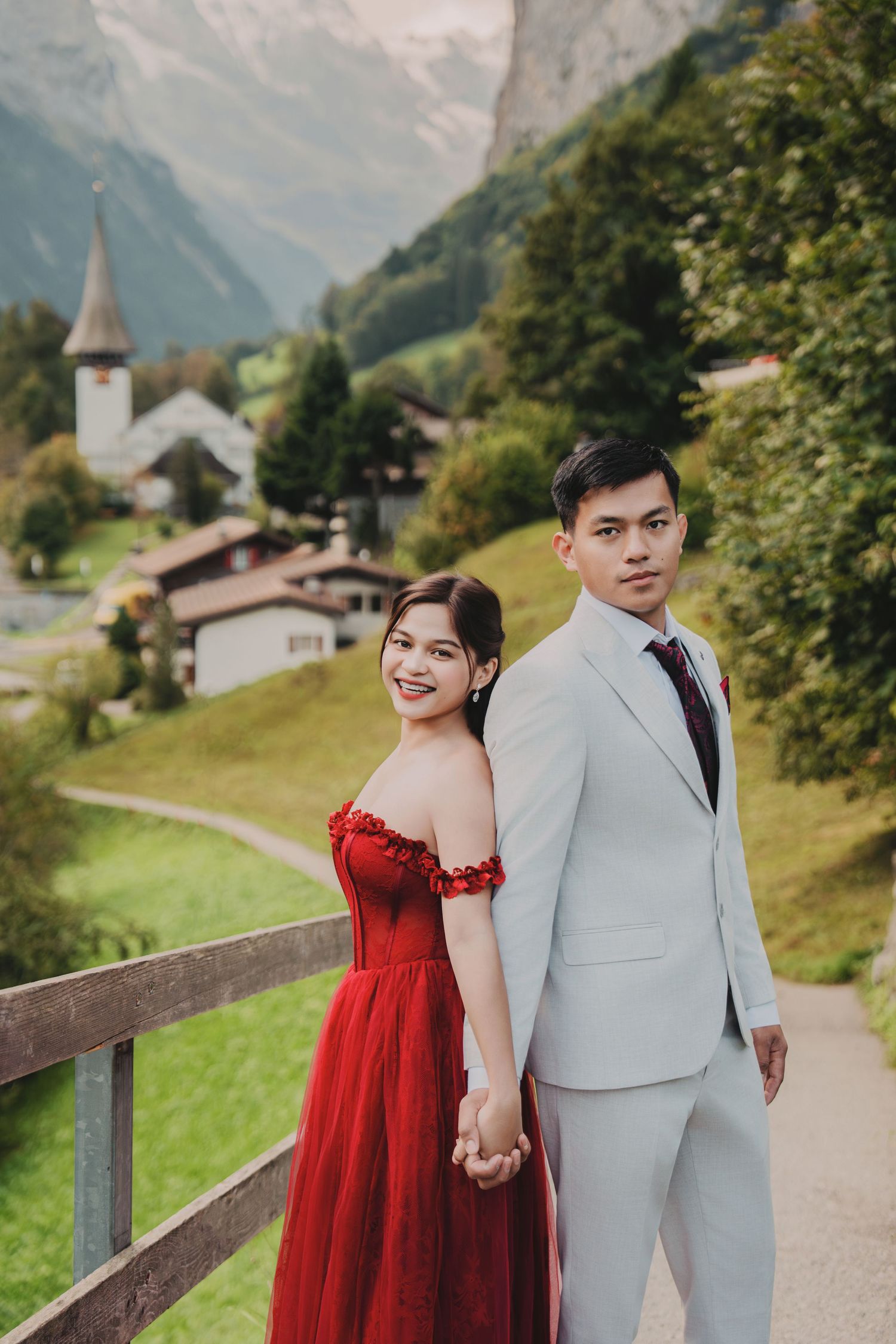 Smiling couple holding hands in Swiss alpine village, woman in red gown and man in grey suit.