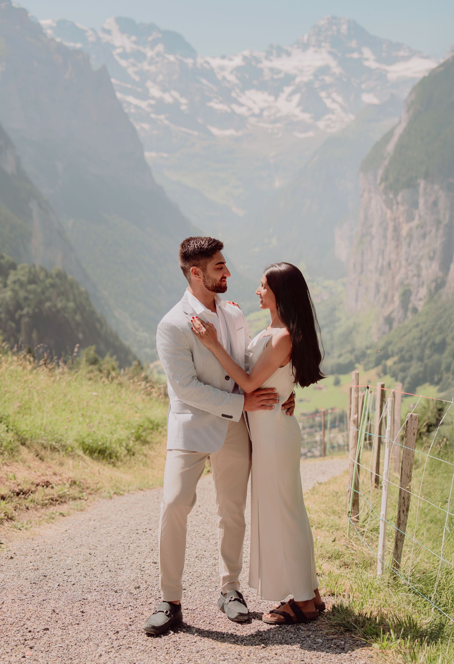 Elegant couple in cream wedding attire embracing on a scenic mountain road with snowy Alps behind.