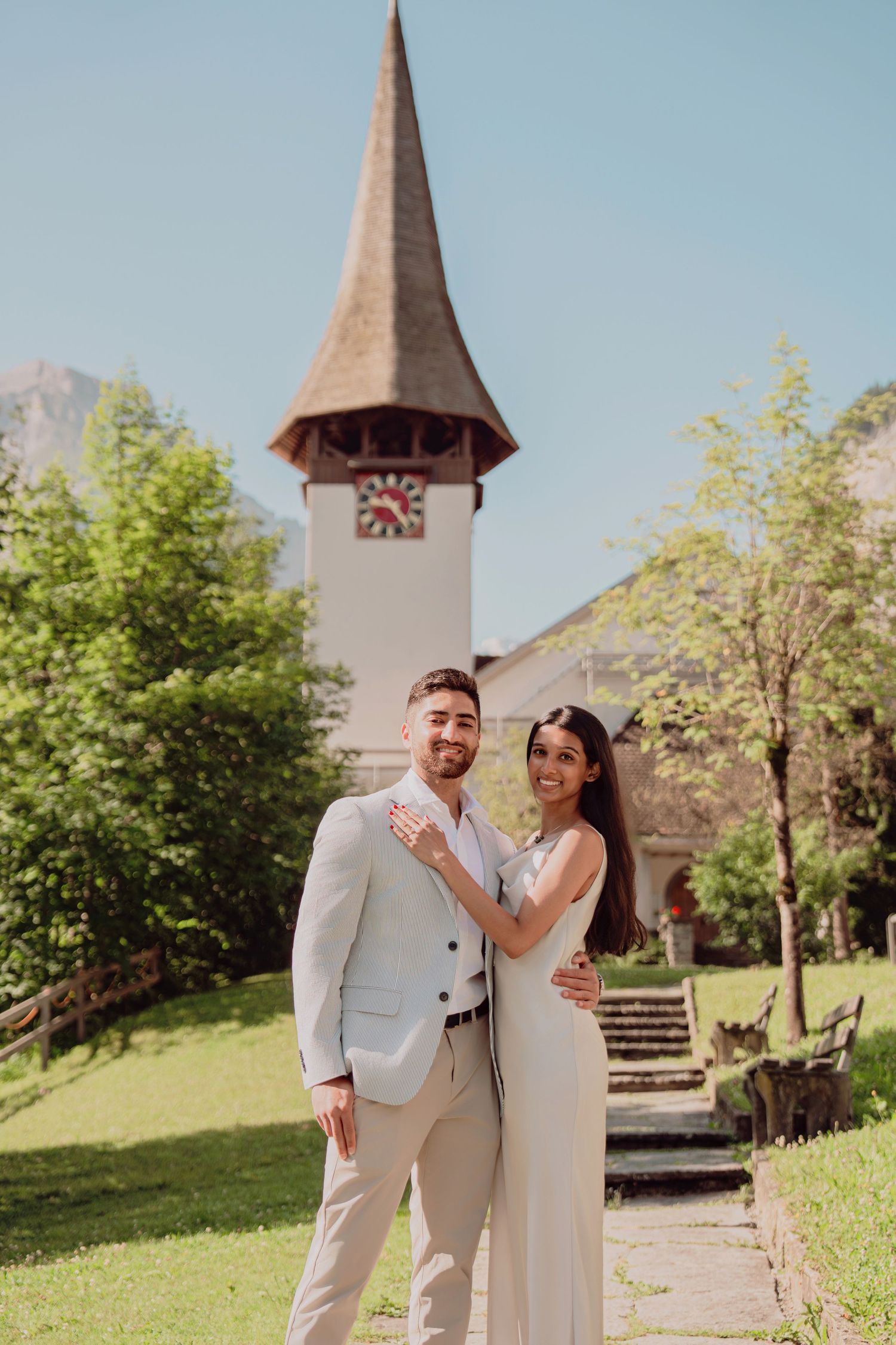Couple poses in front of a charming church with a clock tower in a scenic Alpine village on their wedding day.