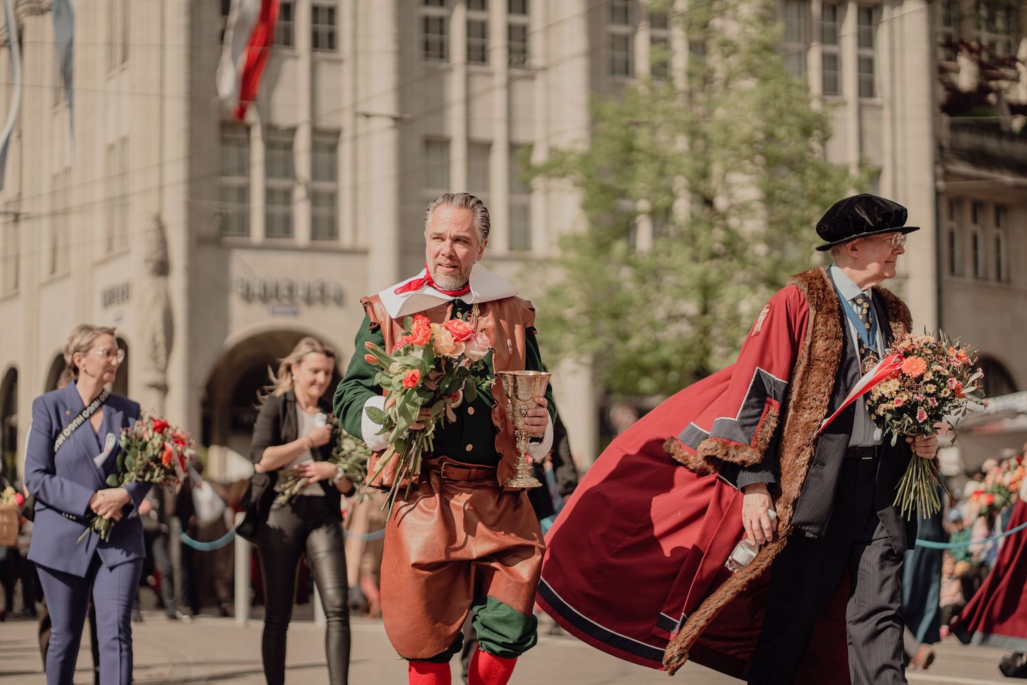 People in Renaissance costumes carry flowers in a festive historical parade through a European city square.