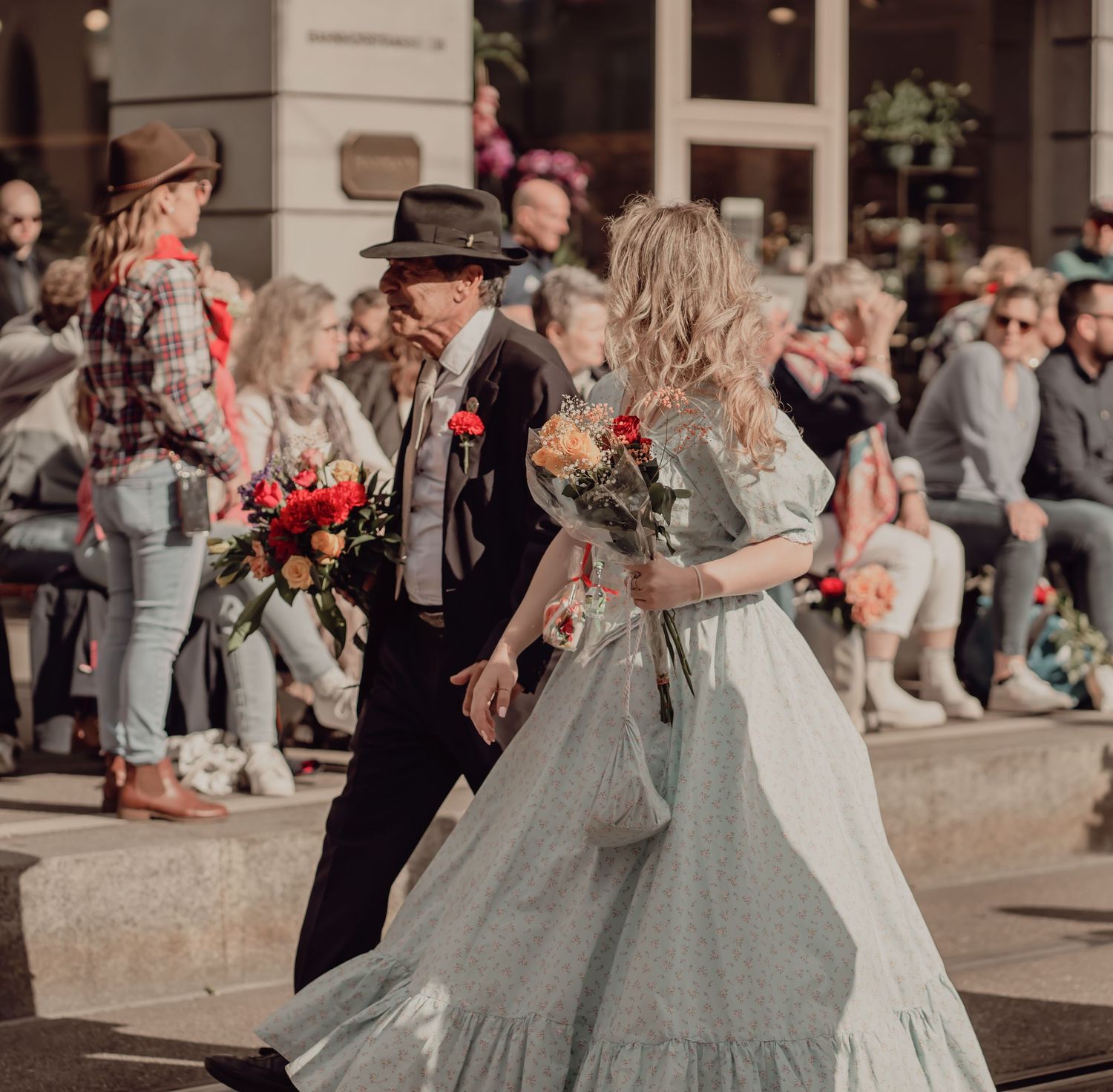 Couple in vintage attire walking with bouquets during an outdoor parade ceremony.