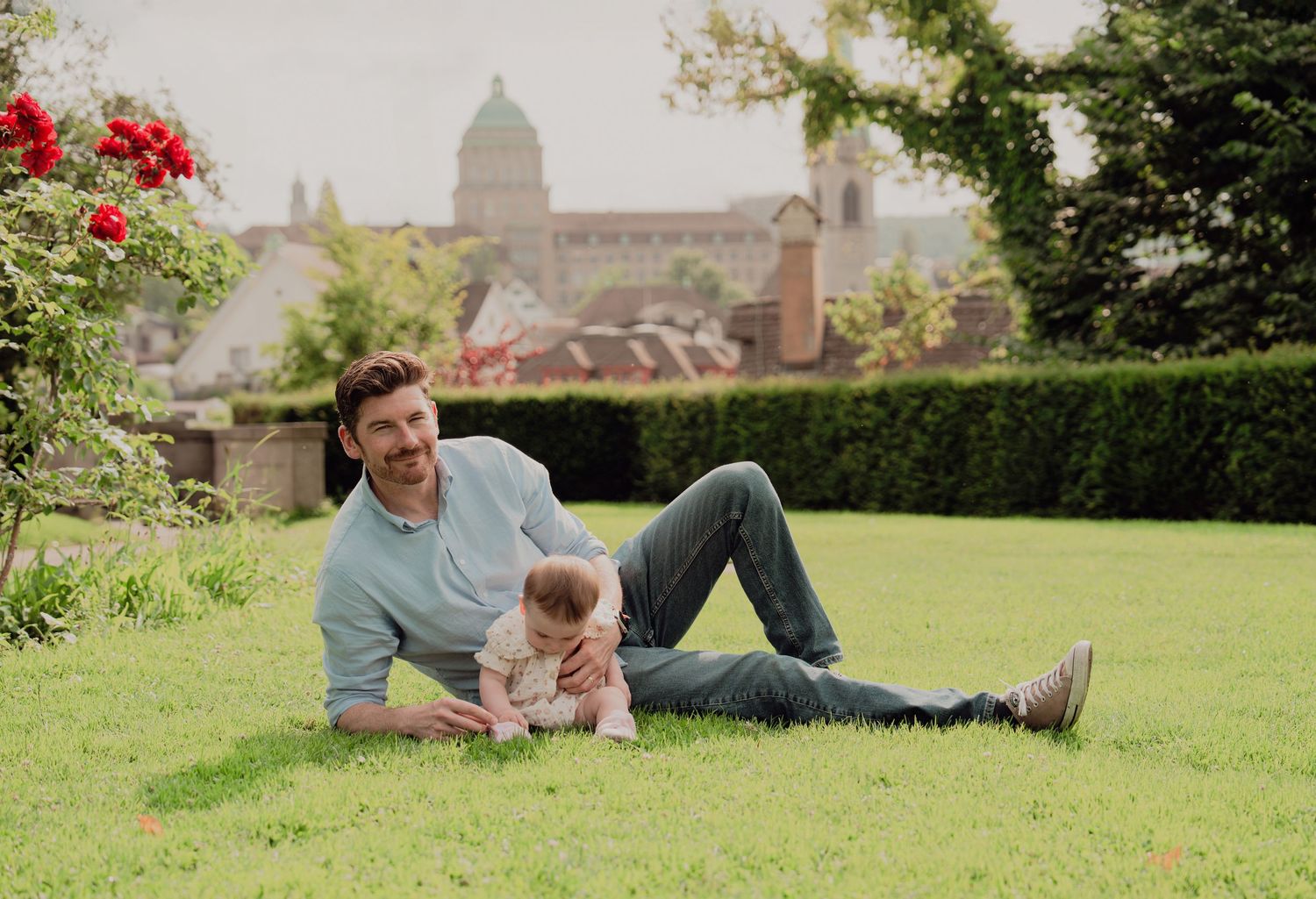 Father in blue shirt lying on green grass with baby in a park with historic buildings in background.
