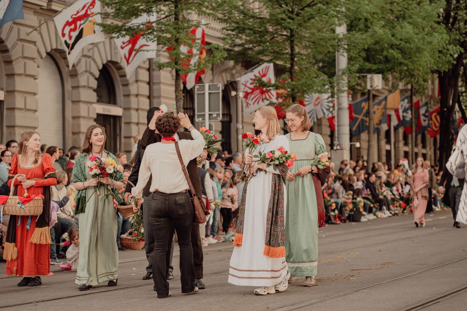 Women in elegant traditional folk costumes carry flower bouquets during a vibrant outdoor street parade.