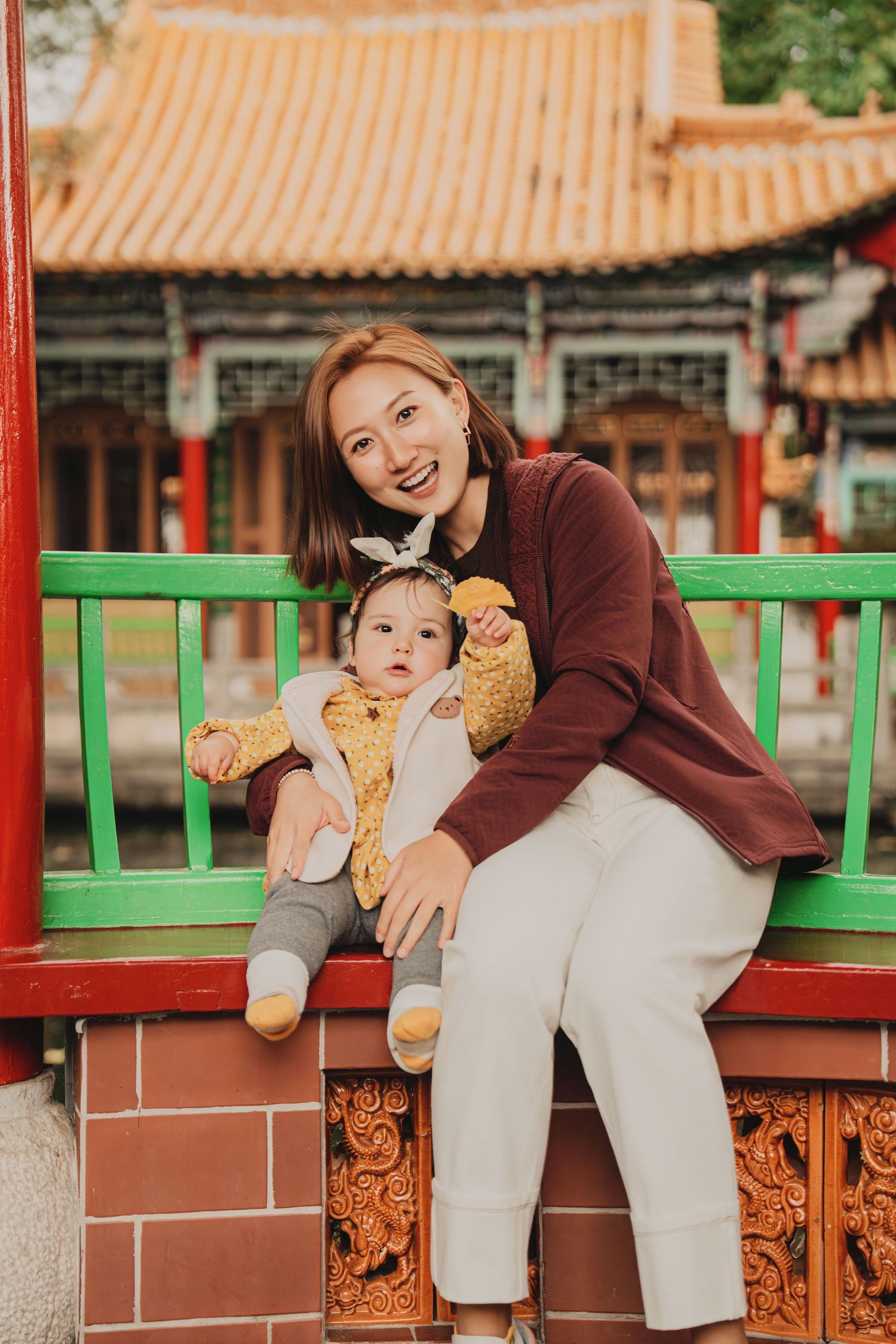 Cheerful Asian mom posing with baby on decorative red Chinese temple balustrade with golden roof.