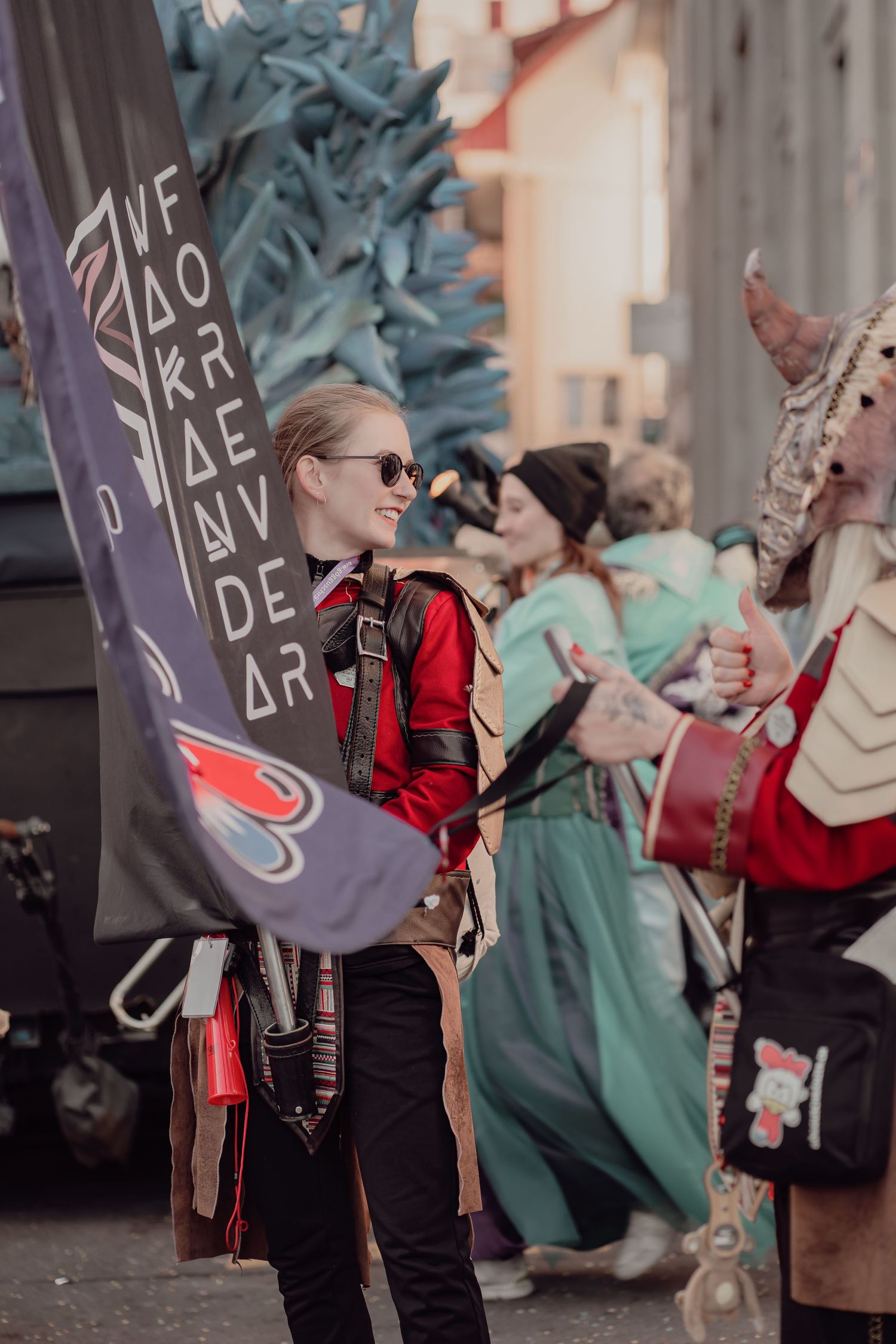 Smiling woman in red costume holds a decorative flag at a fantasy carnival parade.