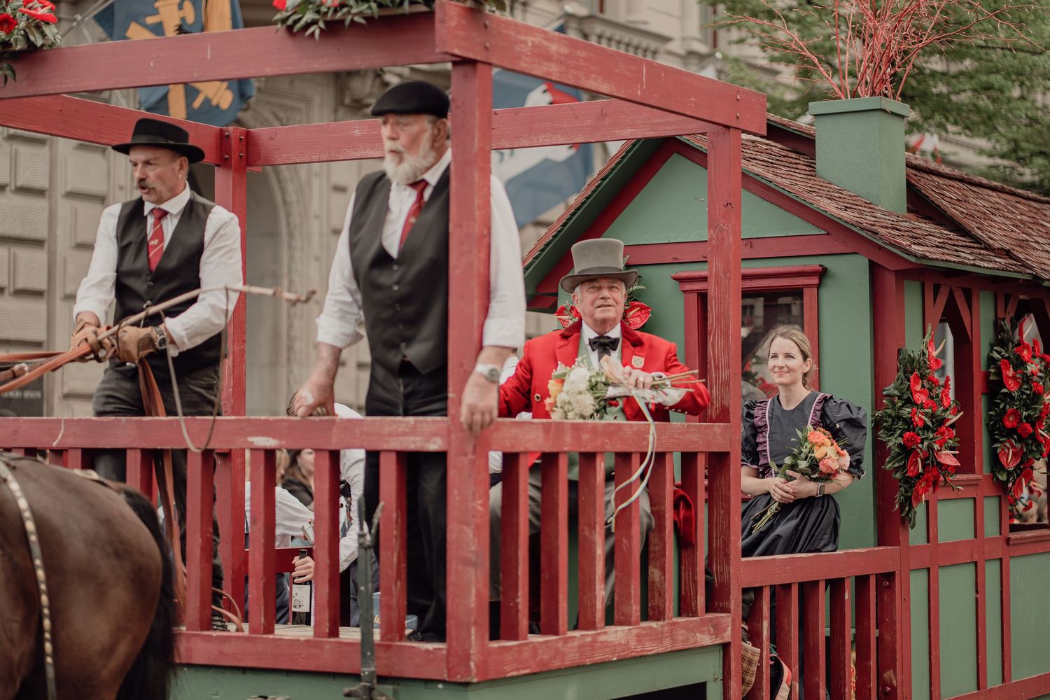 People in Victorian-era costumes wave from a decorated red and green horse-drawn carriage in a parade.