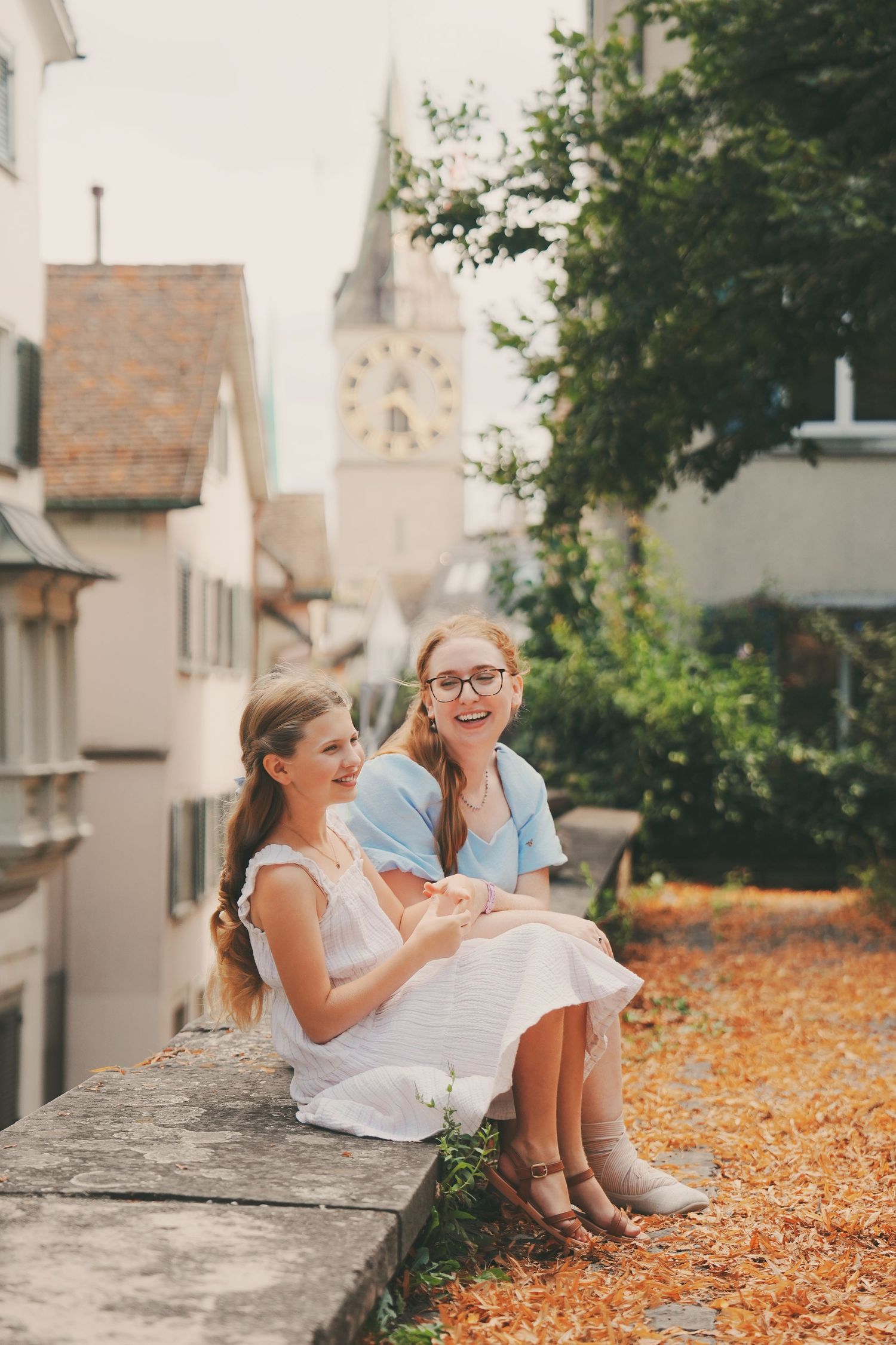 Two females laugh together while sitting on a stone wall with a European church tower in the background.