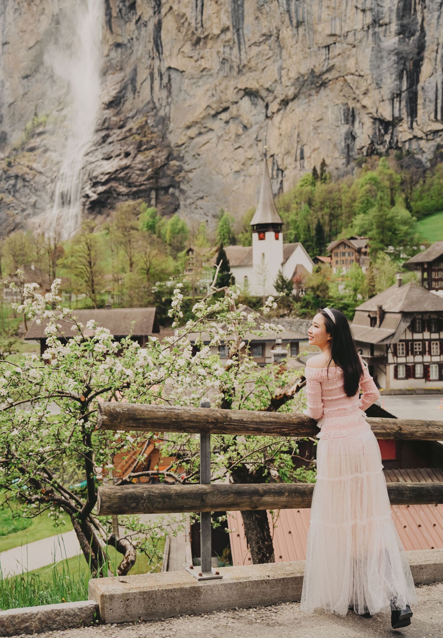 Woman in pink dress admiring Staubbach waterfall and Lauterbrunnen village church amid spring blossoms.