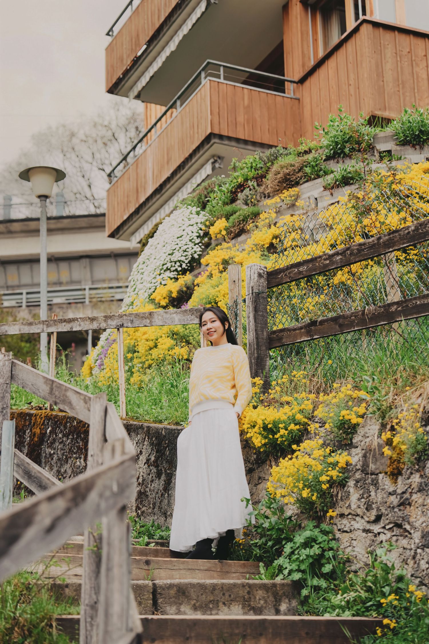 Woman in yellow sweater and white skirt standing on stairs with colorful garden backdrop.