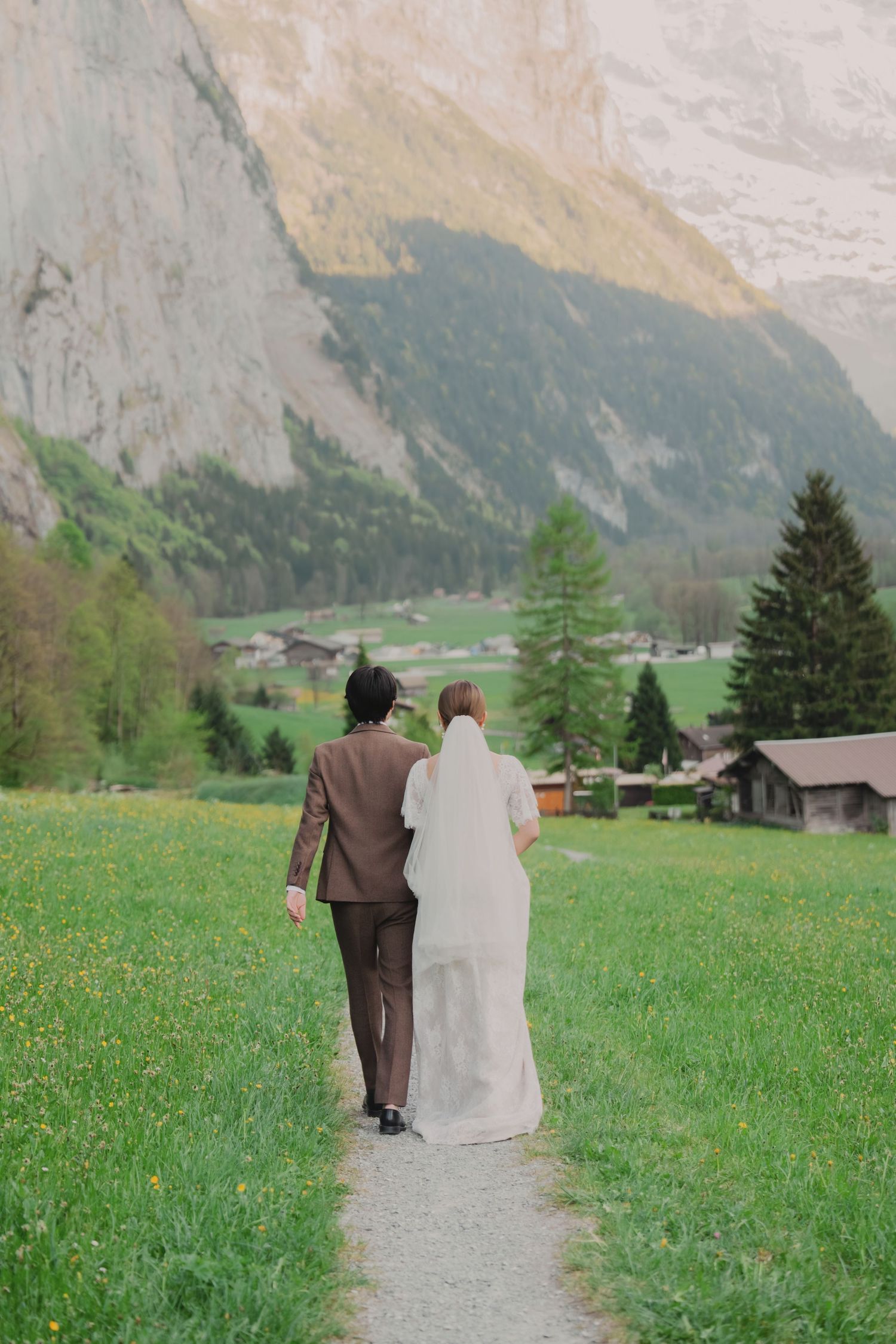 Bride and groom walking hand in hand along a path through green alpine meadows with dramatic Swiss mountains.