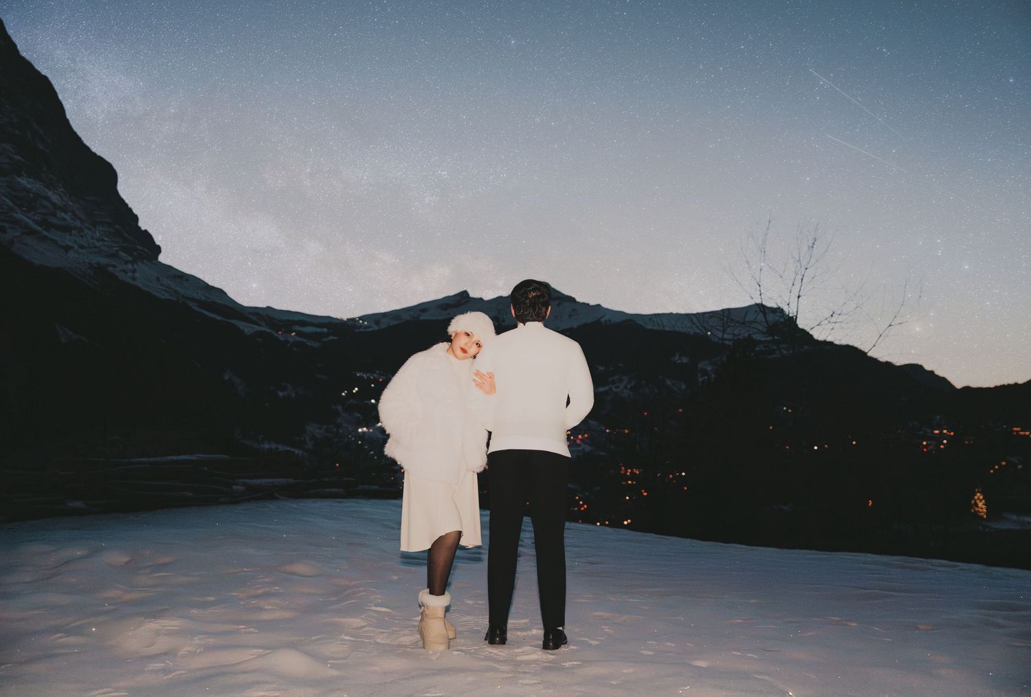 Couple walking hand in hand in white outfits across snowy mountain landscape at dusk.