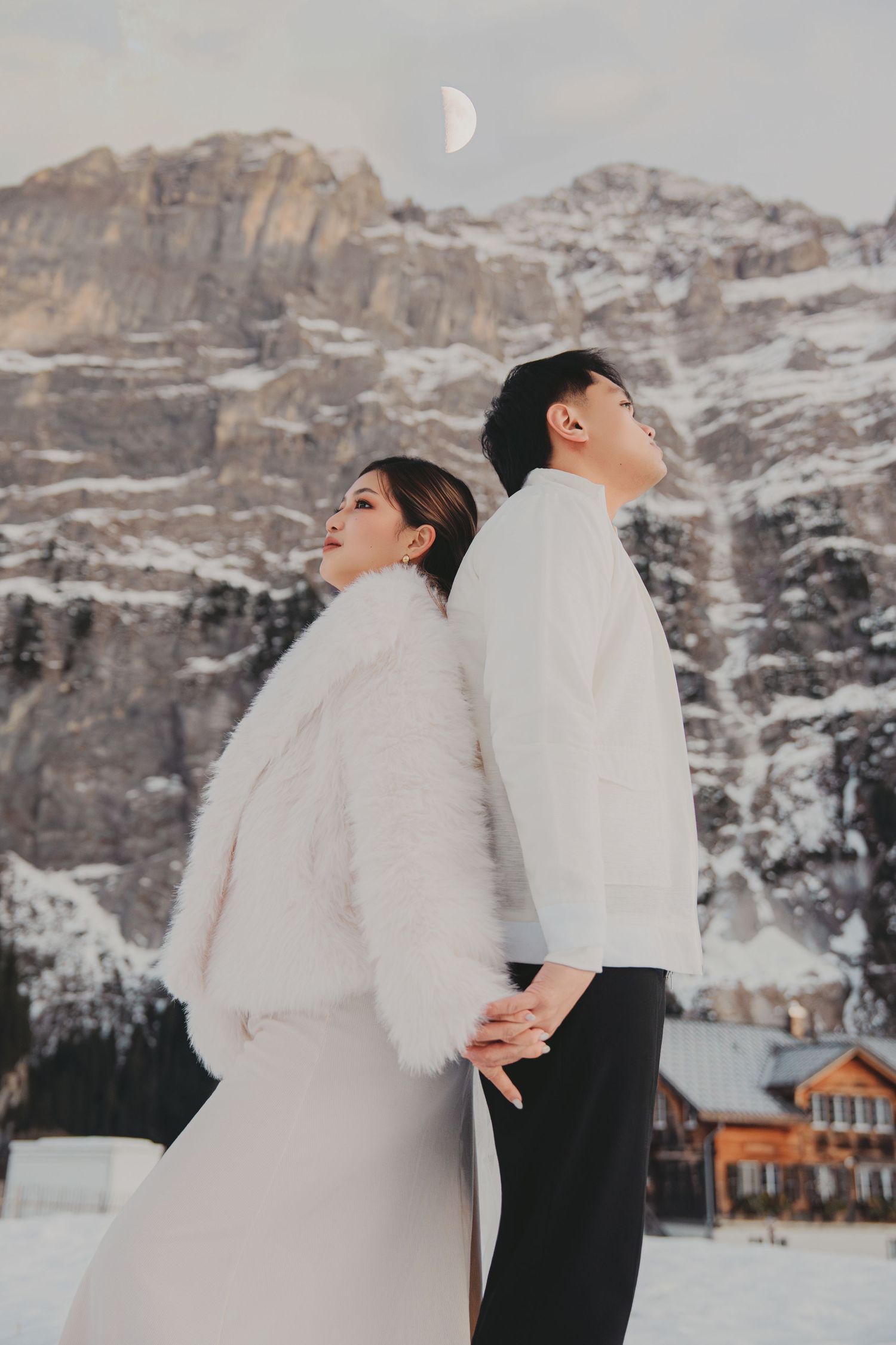 Couple standing back-to-back holding hands in white outfits with snowy Alps and crescent moon above.