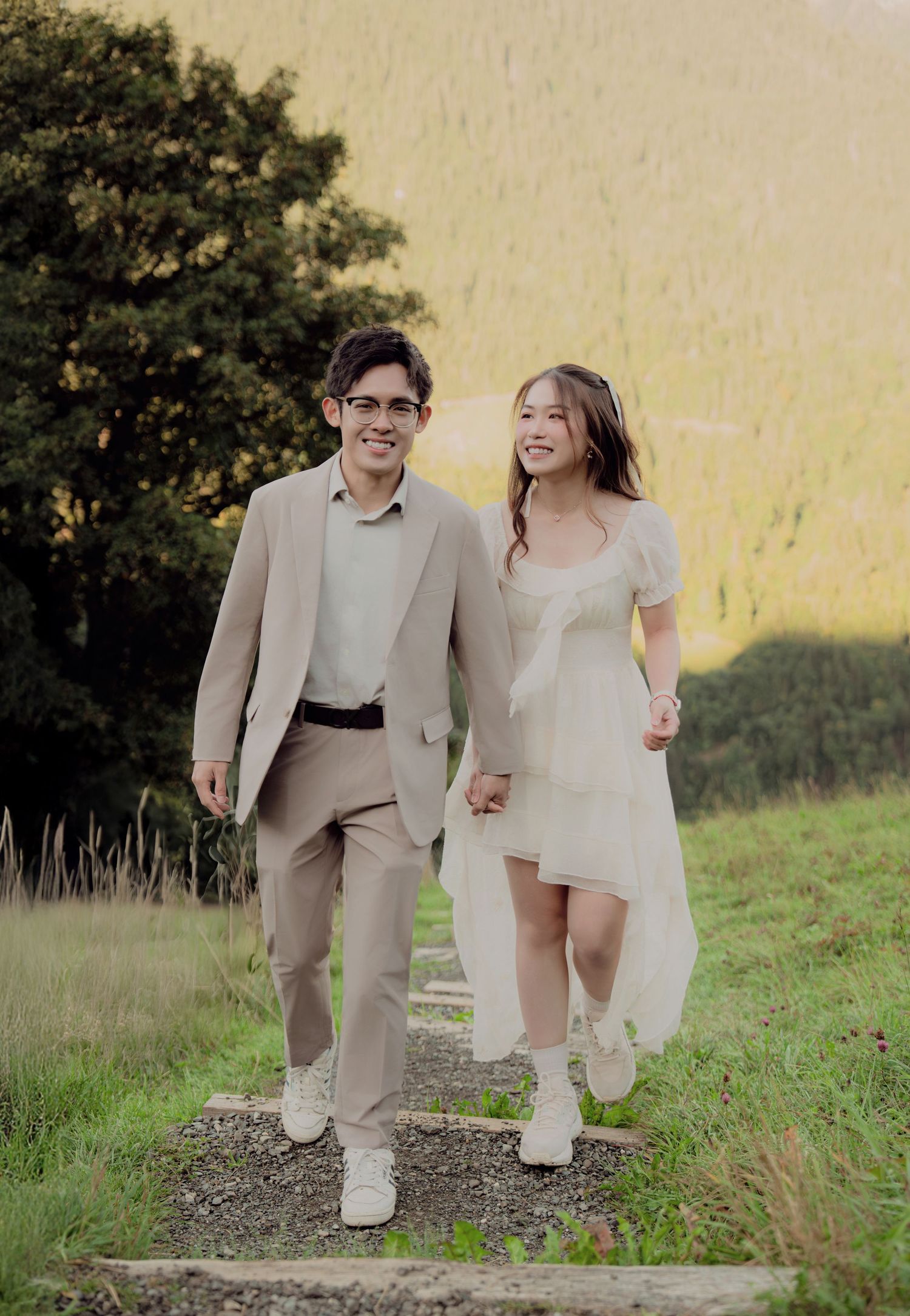 Smiling couple in wedding attire walking hand in hand along a gravel path through a lush green Alpine meadow.