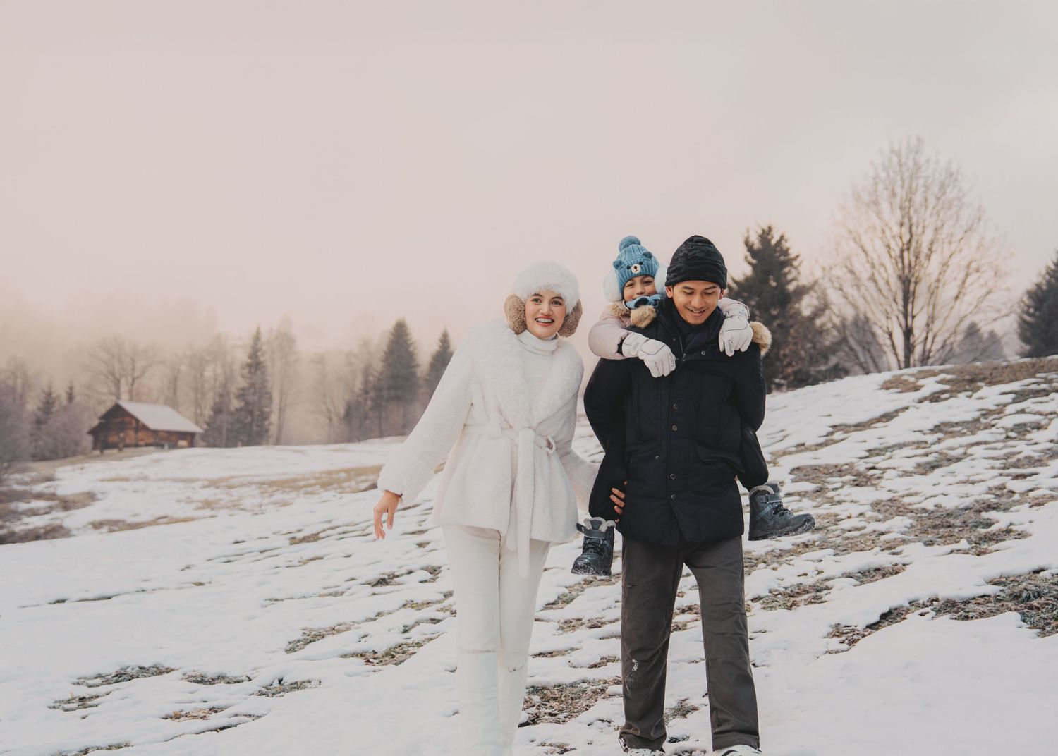 Family laughing and posing on snowy slope with misty pine trees and wooden cabin in winter setting.