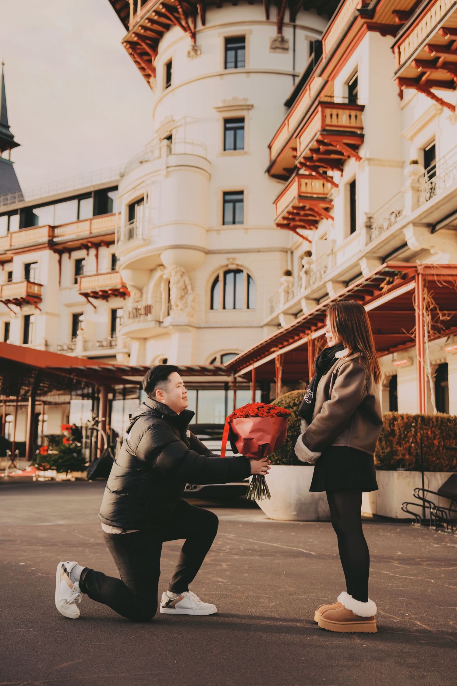 Man on one knee proposing with red roses to woman outside elegant white building.