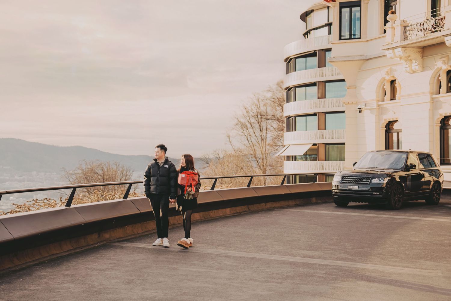 Stylish couple standing together on a scenic terrace with mountain views and a luxury Range Rover.