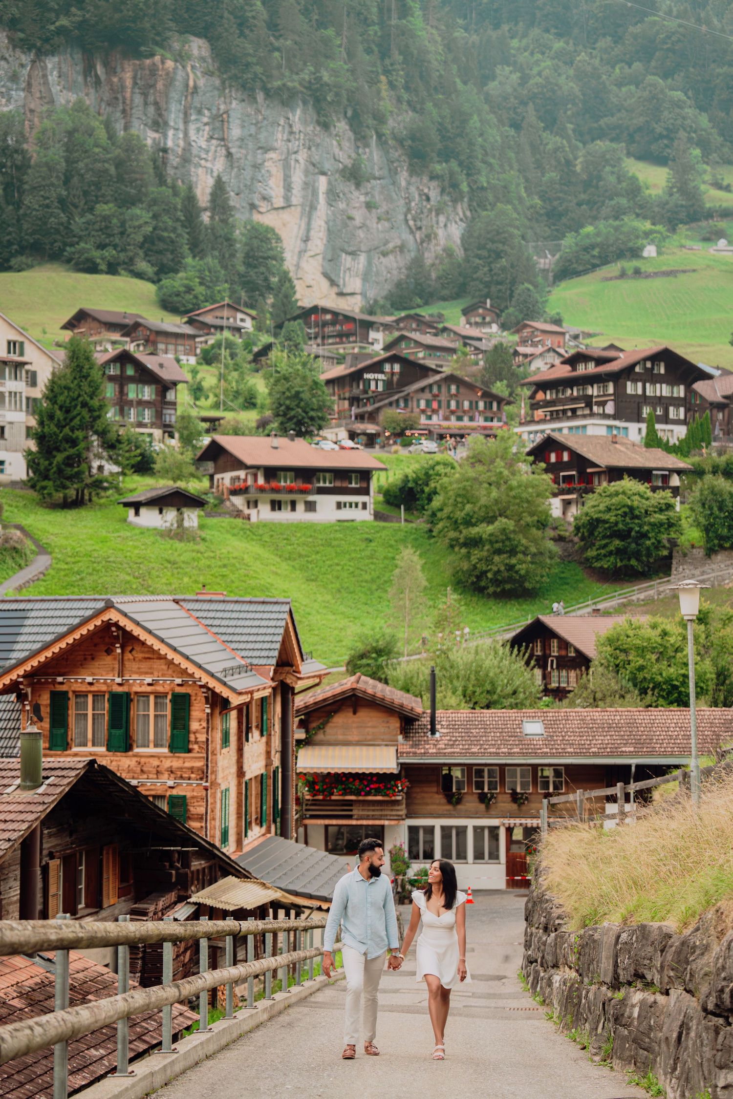 Couple strolling hand in hand through a scenic Swiss Alpine village with traditional wooden chalets on hillside.