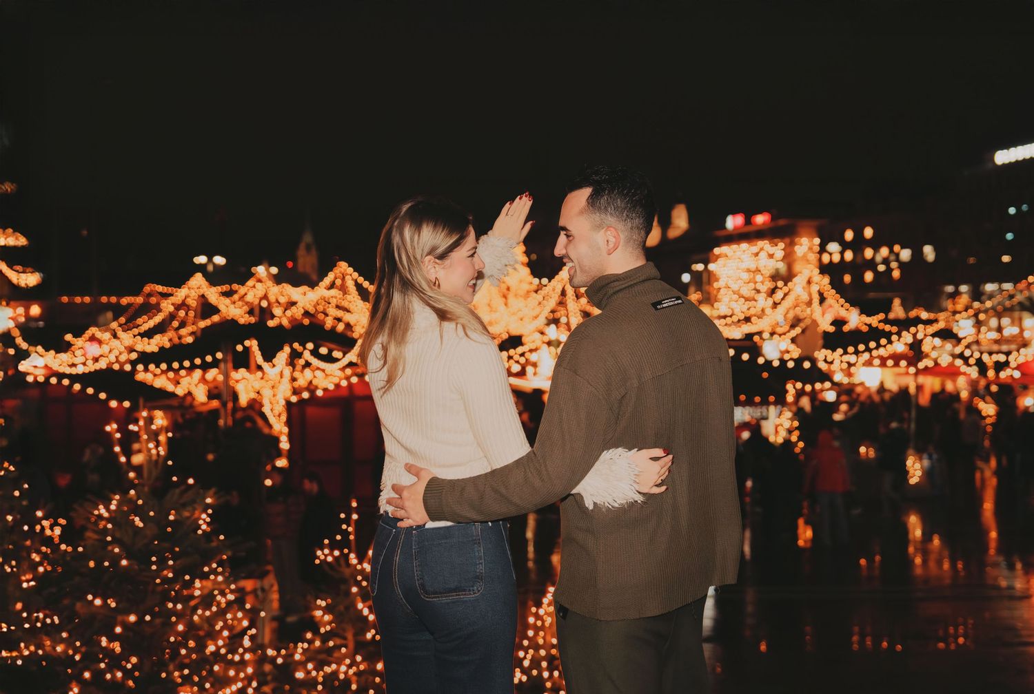 Couple smiling and dancing together at a vibrant Christmas market with warm golden lights at night.
