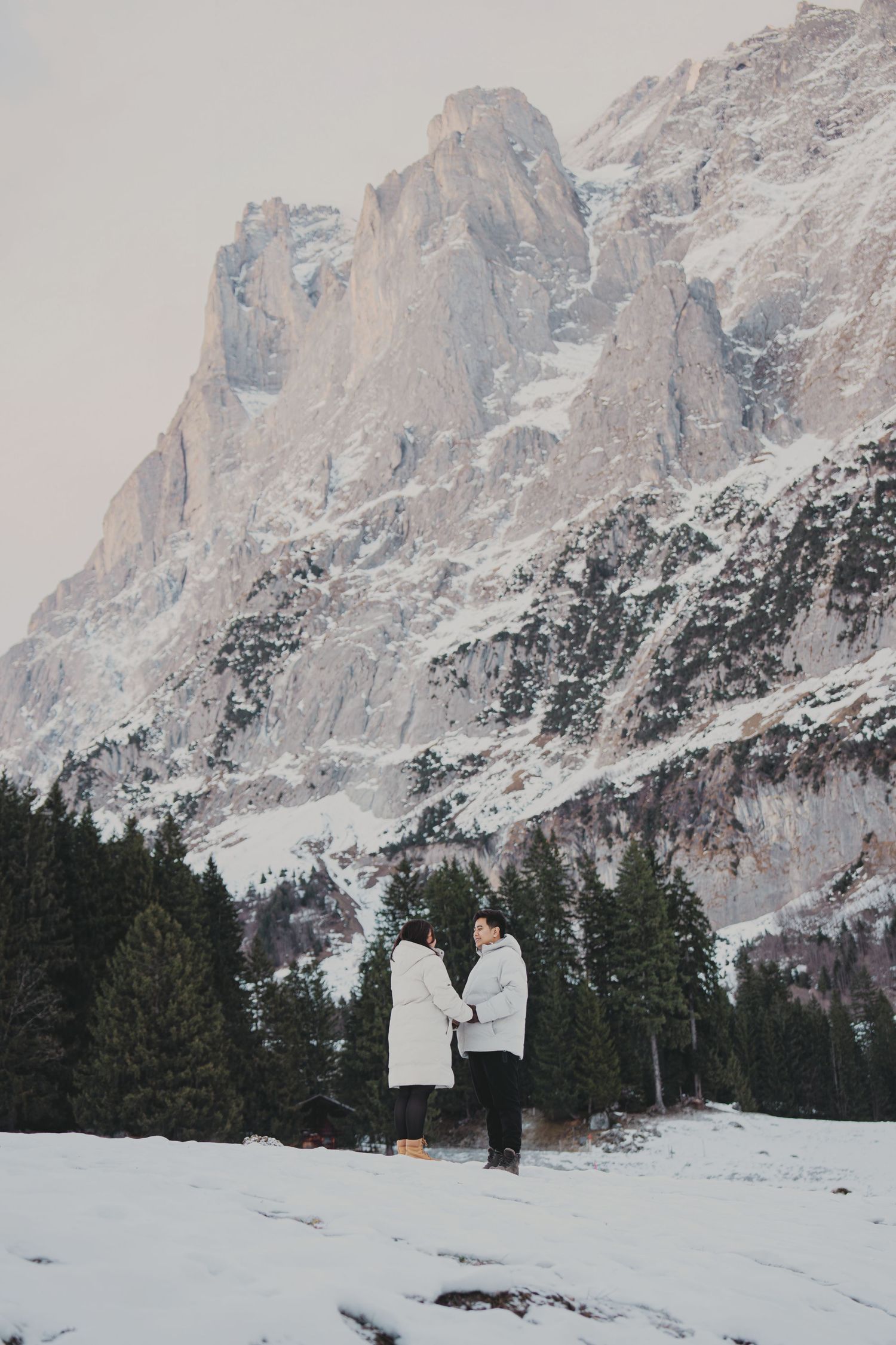 Couple holding hands facing each other on snowy ground with dramatic snow-covered mountain peaks behind.