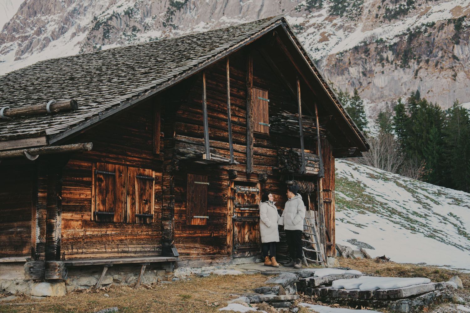 Couple in matching white coats standing at entrance of rustic alpine log cabin in winter mountains.