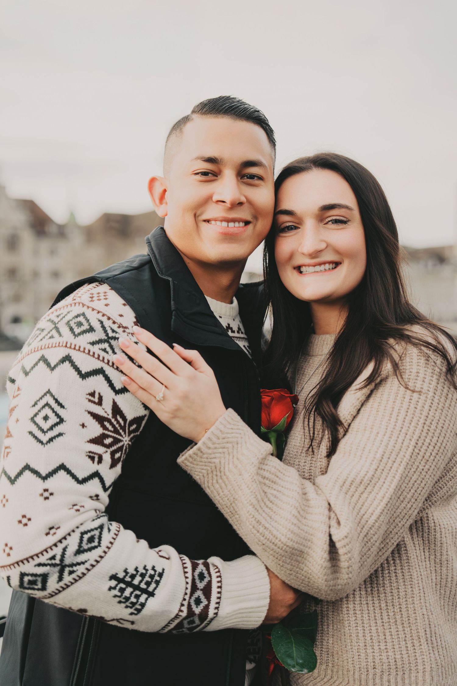 Happy newly engaged couple smiling and holding red rose with Zurich skyline behind.
