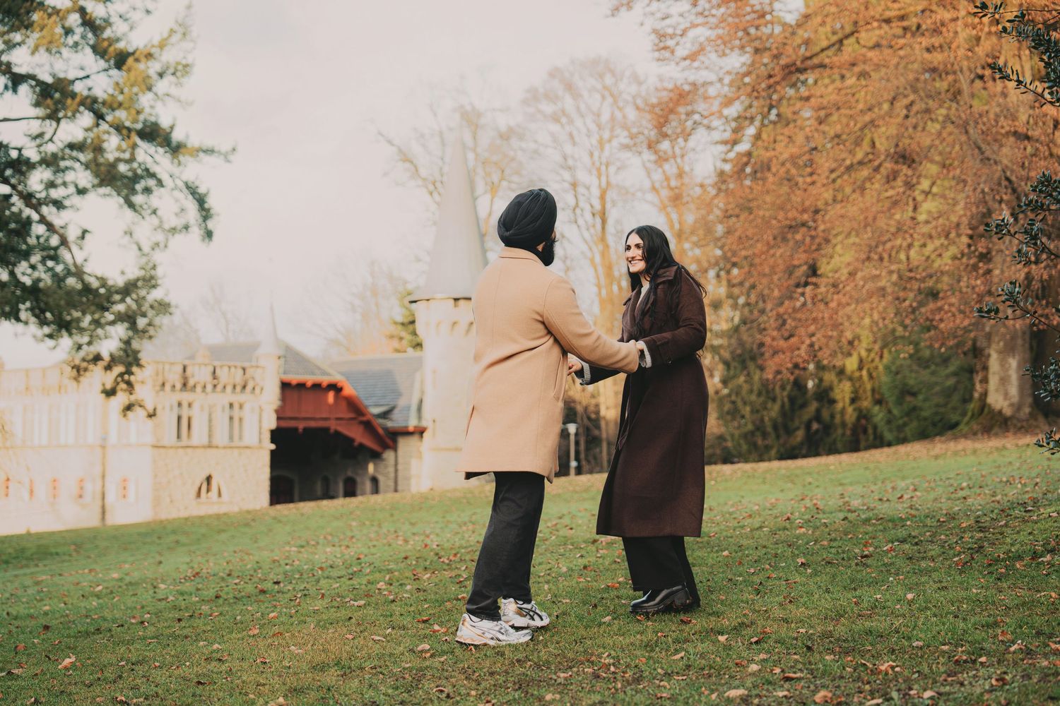 Couple holding hands facing each other on a grassy lawn with colorful autumn trees nearby.