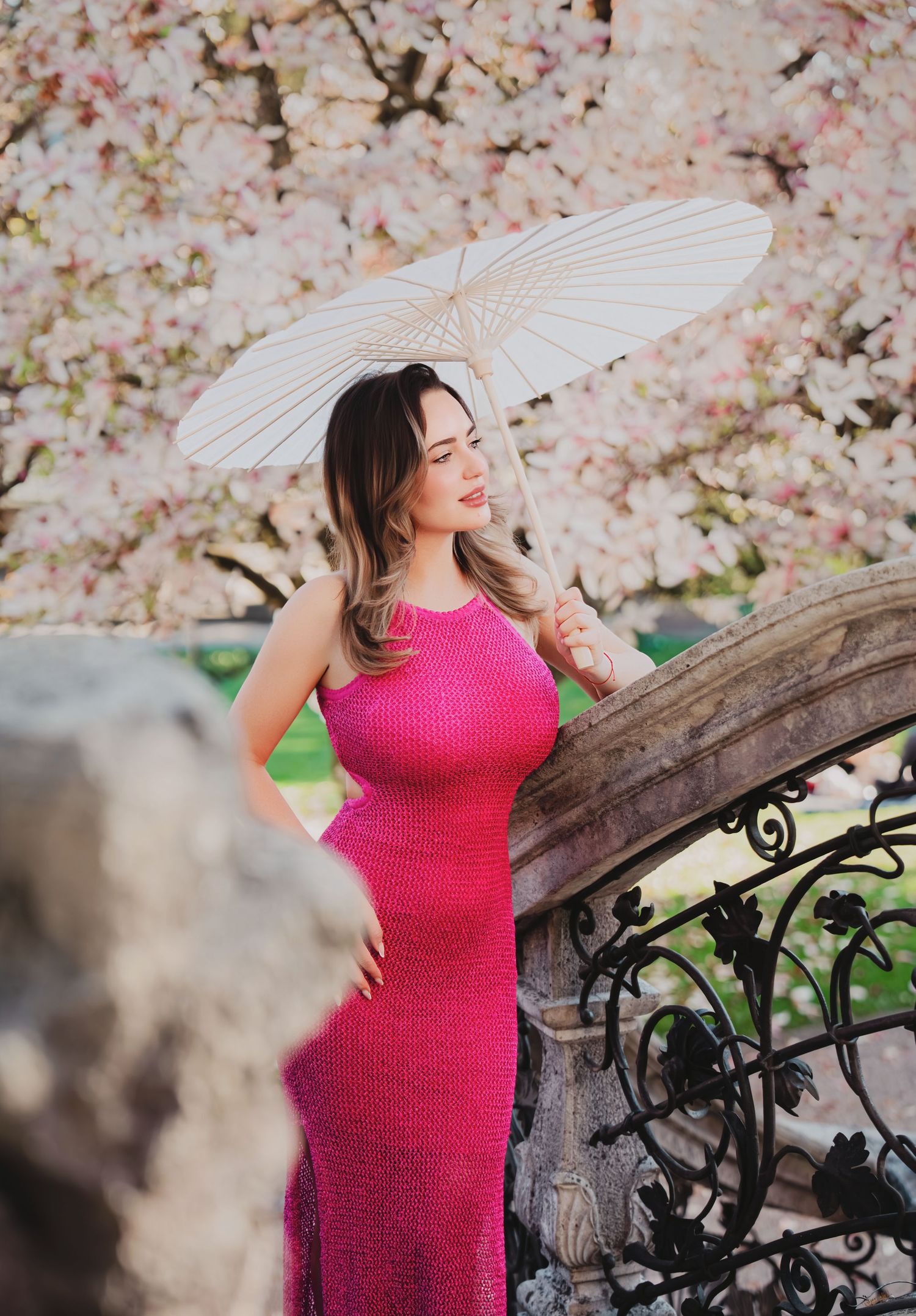 Woman in pink knit dress holding white parasol under cherry blossom trees on stone staircase.