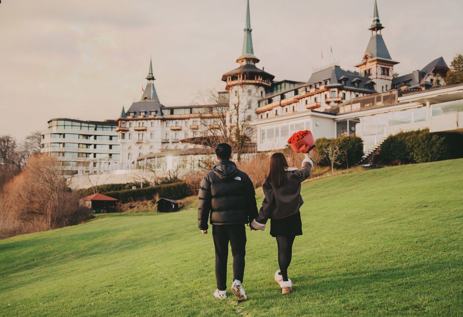Couple walking hand in hand toward grand European castle, woman holding red flowers.