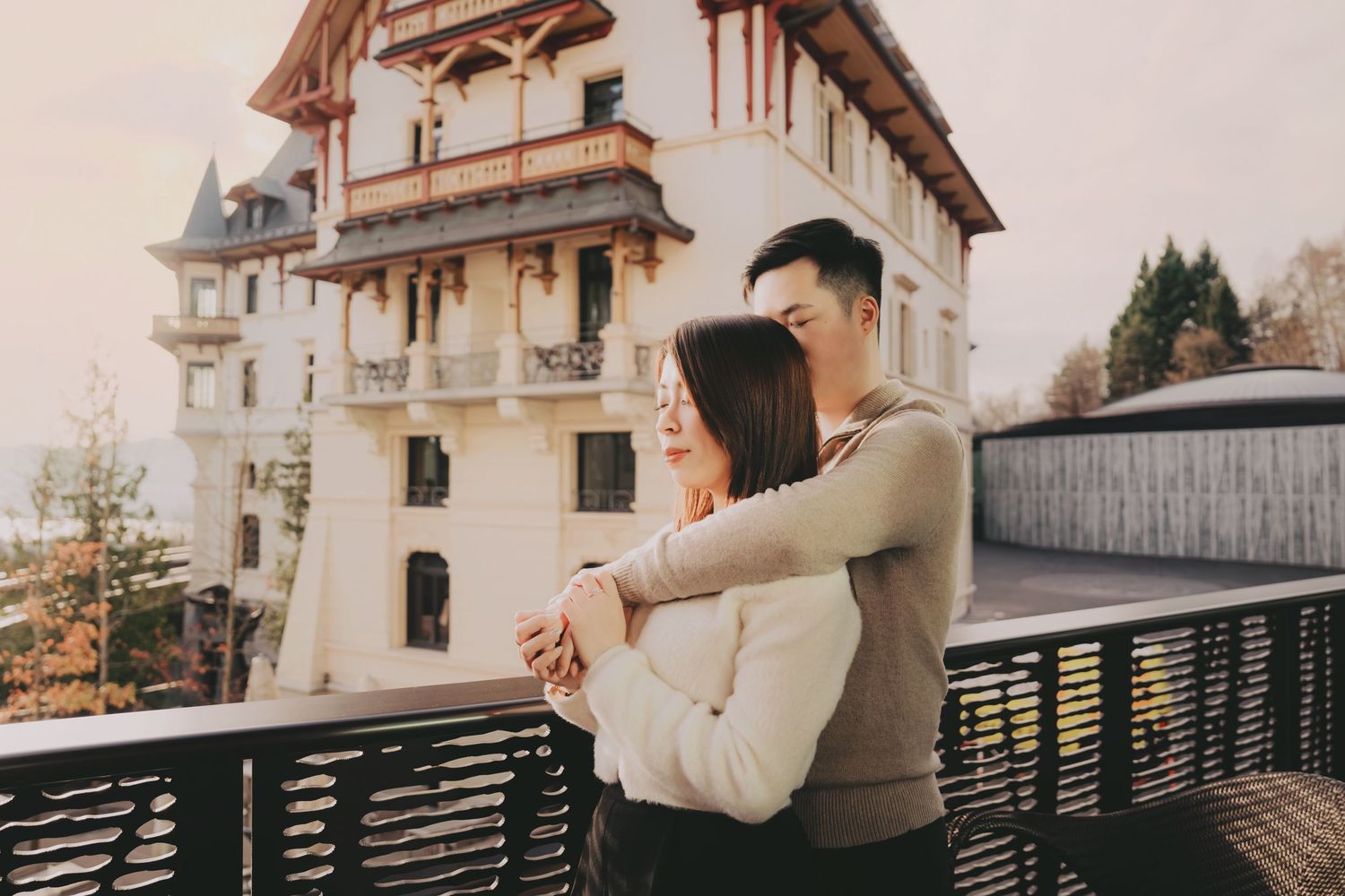 Asian couple sharing a romantic embrace on a balcony with a charming vintage building backdrop.