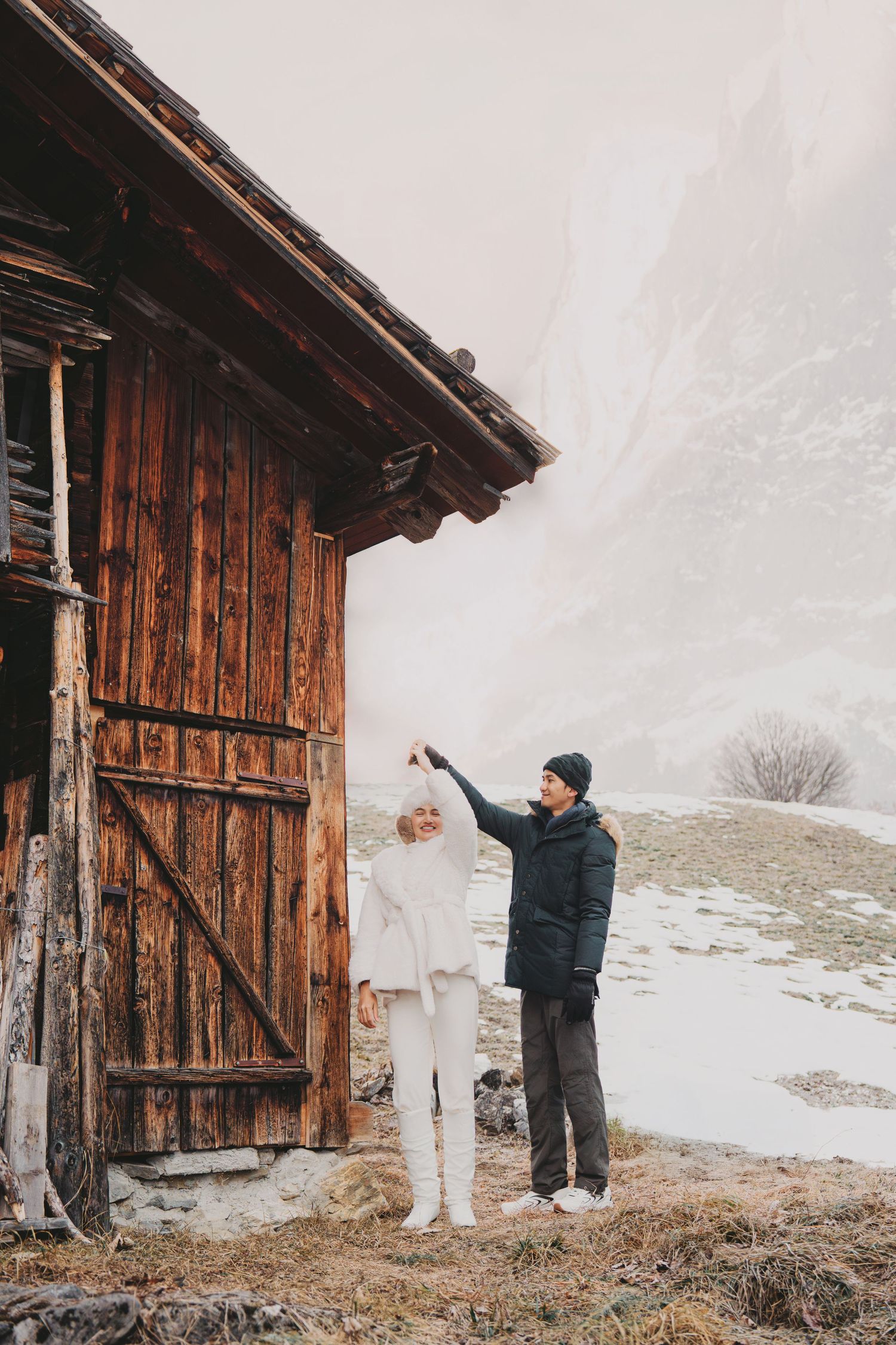 Couple dancing playfully outside a rustic wooden barn in a snowy mountain setting.